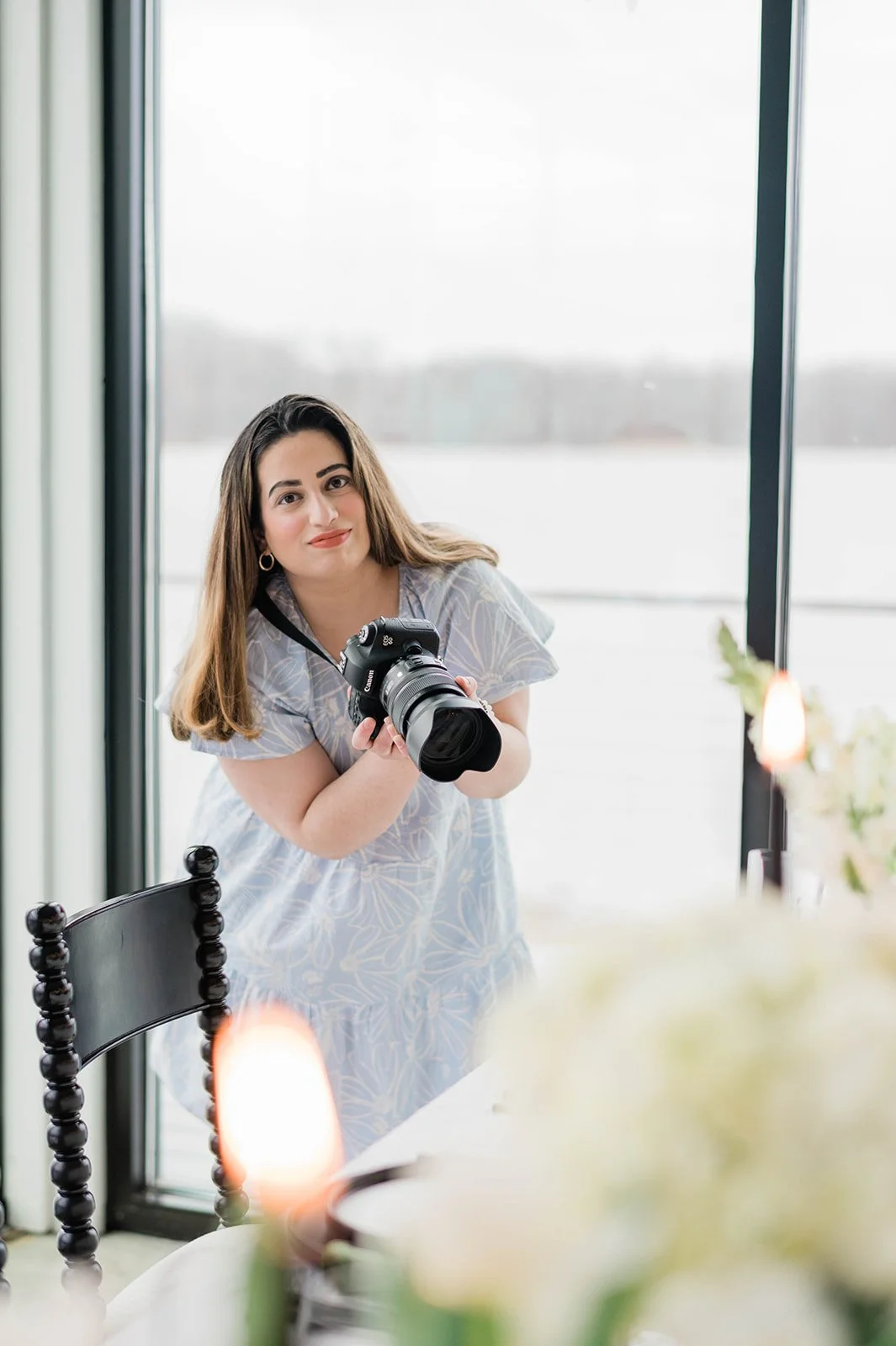 A young woman with long brown hair holding a professional camera, standing by a table with soft lighting and looking towards the camera in a bright, modern space with large windows showing a snowy outdoor scene.