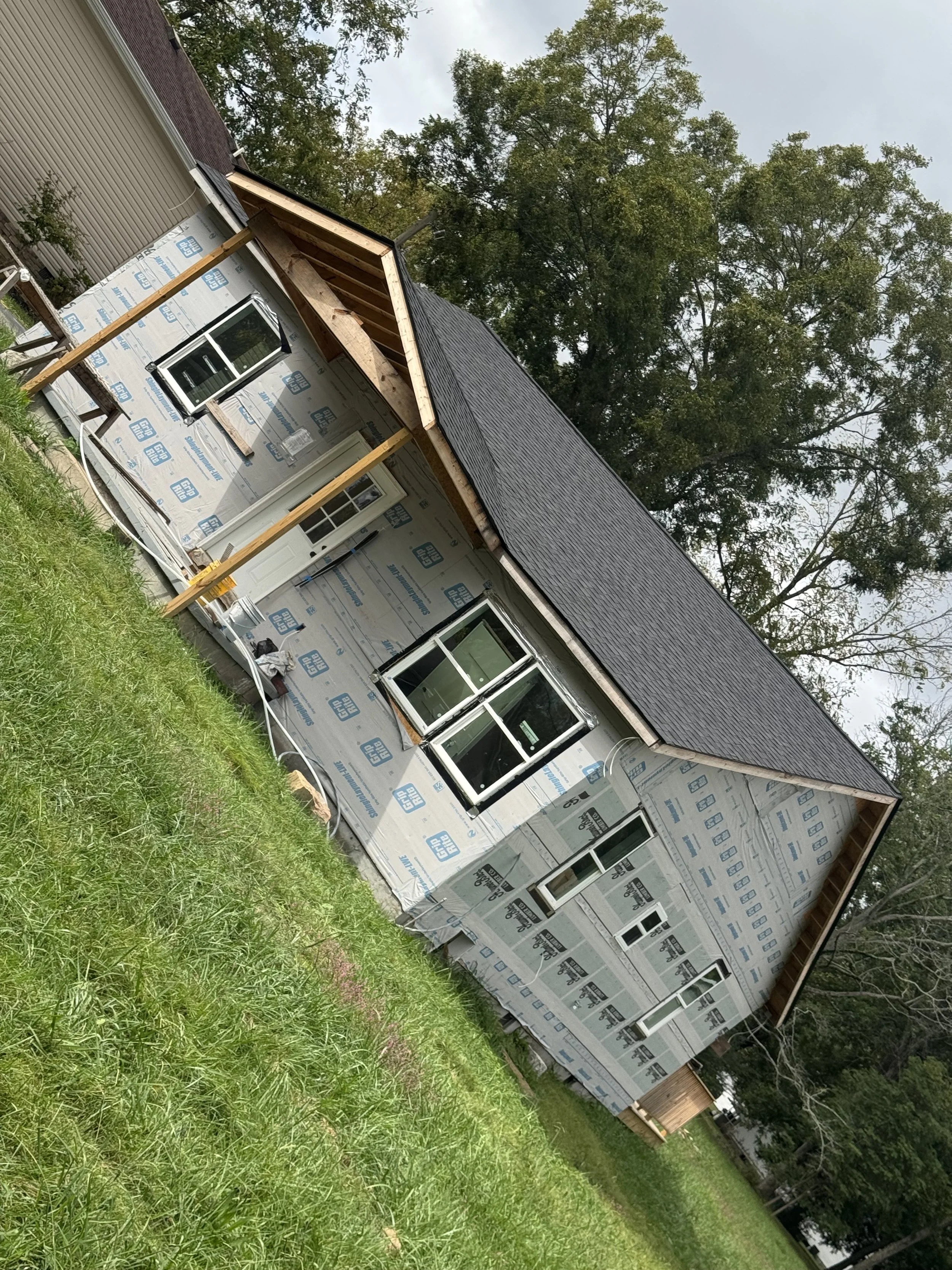 A house under construction with a black shingle roof and wrapped exterior walls, and multiple windows installed, situated on a grassy yard with trees in the background.