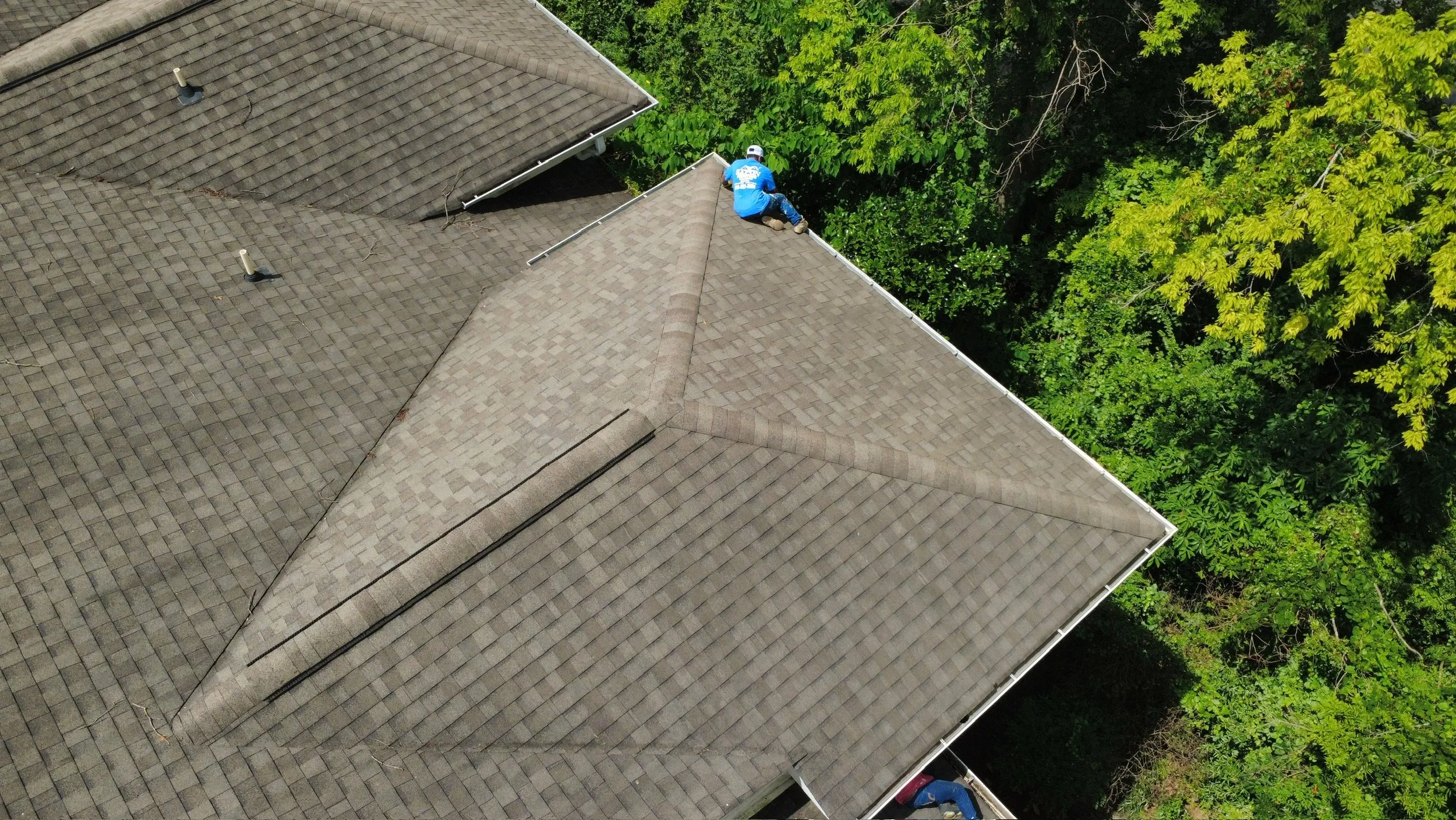 An aerial view of a house roof with a worker on top, surrounded by trees.