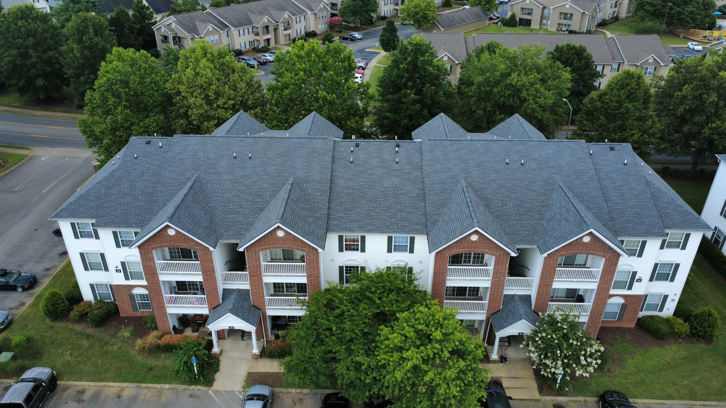 Aerial view of a three-story apartment complex with a brick and white siding exterior, surrounded by parking lots, trees, and neighboring residential buildings.
