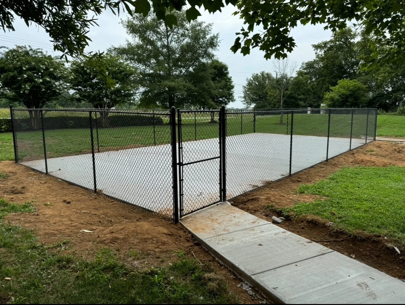 A fenced-in outdoor tennis court with a concrete surface, surrounded by grass and trees in a park-like setting.