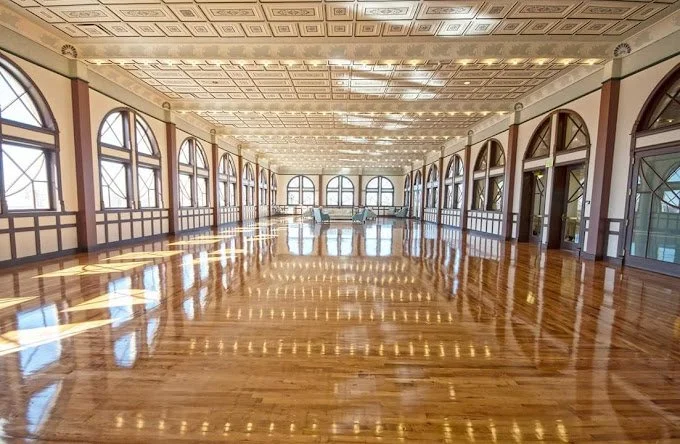 Large, empty ballroom with polished wooden floors, high ceilings, and large arched windows allowing natural light.