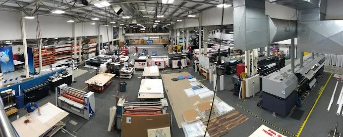 Manufacturing or printing facility with machinery, work tables, and materials organized on shelves and floors.