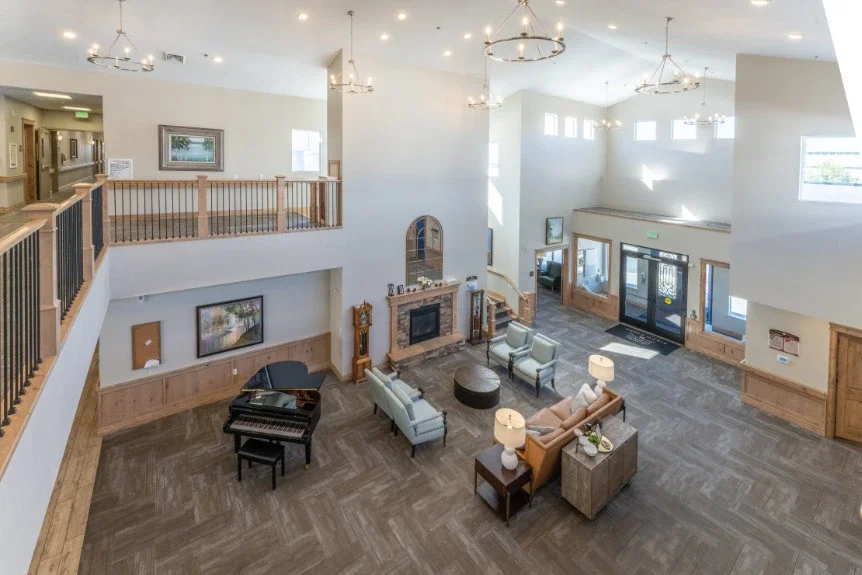 Spacious hotel lobby with high ceiling, modern chandeliers, seating area with sofas and armchairs, a fireplace, and a grand piano, featuring light-colored walls and wooden accents.
