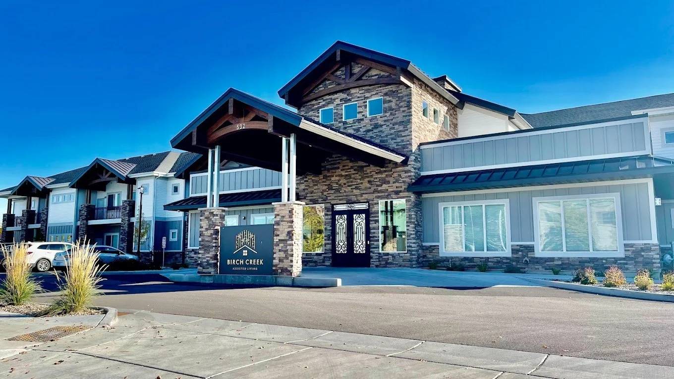 Modern apartment complex with stone and siding exterior, parking lot, and a sign that reads "Birch Creek Assisted Living" under a clear blue sky.