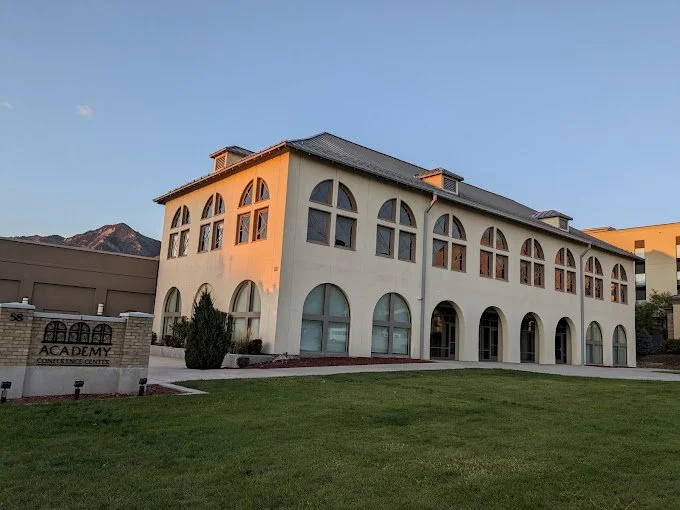 A large, white, multi-story building with arched windows and a dark roof, surrounded by a well-maintained lawn, with a sign that reads "ACADEMY CONCRETE DESIGN".