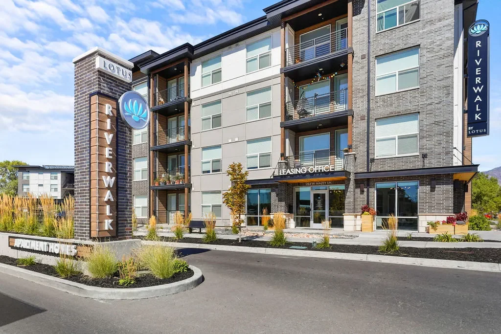 Exterior view of a modern apartment complex named 'Riverwalk' with a leasing office, landscaped surroundings, and signs indicating 'Apartments for Rent' and 'Leasing Office'.