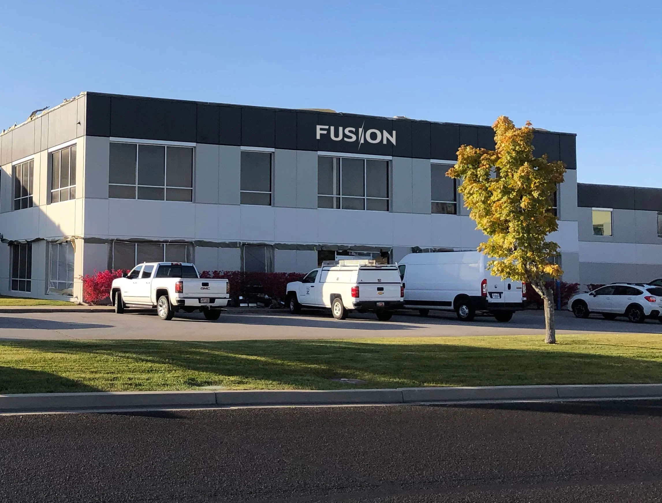 Modern office building with a black and white exterior, large windows, a sign that reads 'FUSION' on the top, parked cars in front, and a tree with yellow-green leaves on a clear day.