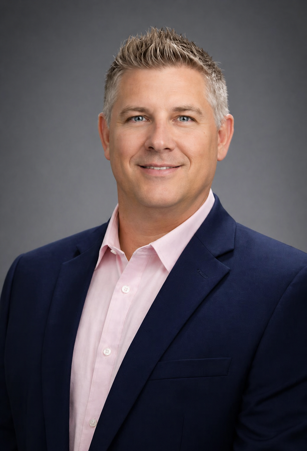 Professional headshot of Patrick Thompson, AWMA,  wearing a navy blazer and a light pink dress shirt, smiling against a gray background.