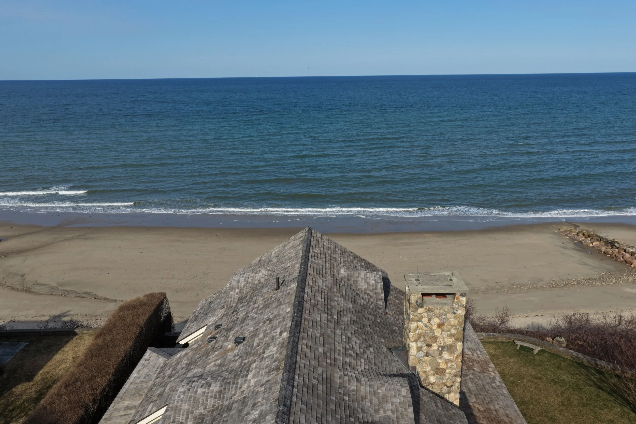 View from an elevated position showing the roof of a house, sandy beach, and ocean with waves and a clear sky in the background.