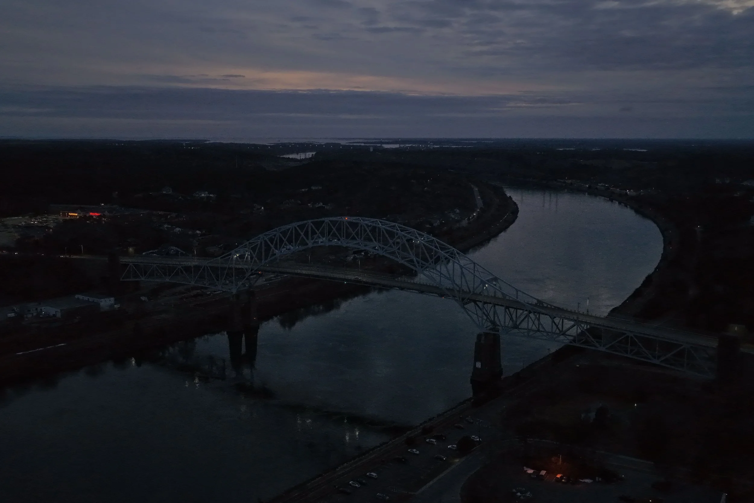 Aerial view of a large steel arch bridge crossing over a wide river at dusk, with a cloudy sky overhead.
