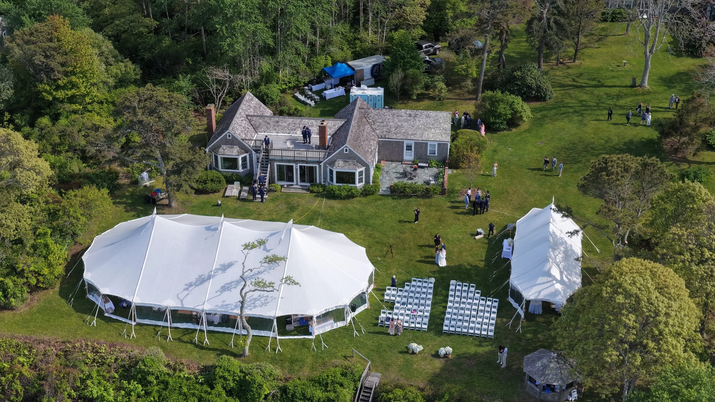 Aerial view of an outdoor wedding reception with a house, large white tent, seating area, and guests on a lush green lawn surrounded by trees.