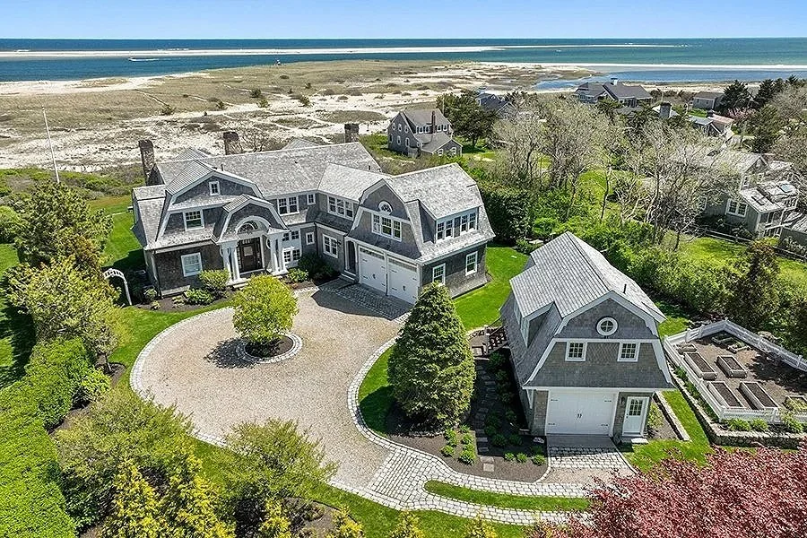 A large seaside home with gray shingles, a round driveway, and lush green landscaping, located near the beach and ocean.