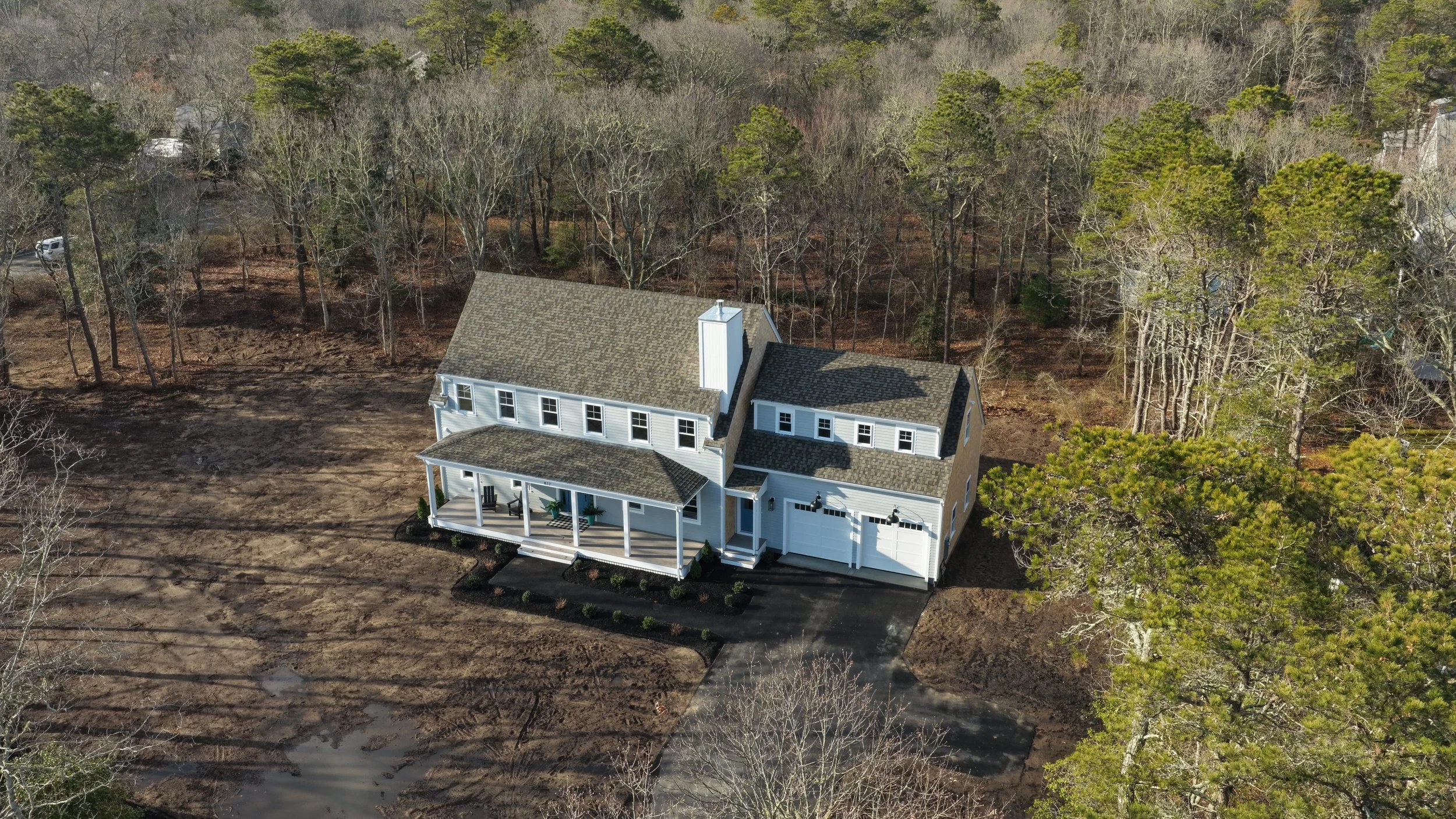 A two-story house with a front porch surrounded by trees, some with leaves and others without, indicating a season transition, in a wooded area.