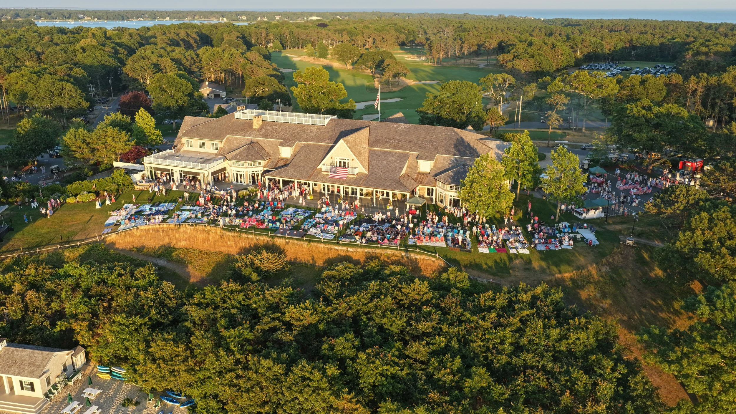 Aerial view of a clubhouse and grounds at a golf course with a party or event taking place outside on the grassy area, surrounded by trees, and with a parking lot and water in the background.