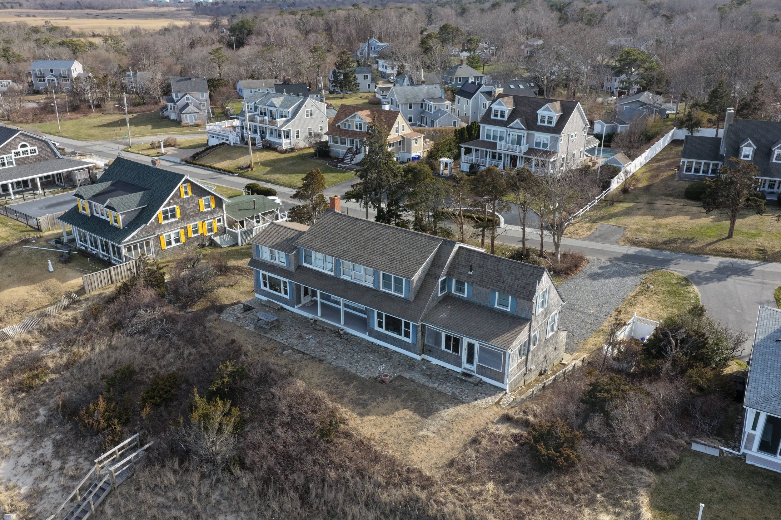 Aerial view of a coastal residential neighborhood with multiple houses, some with porches and yellow shutters, surrounded by trees and lawns.