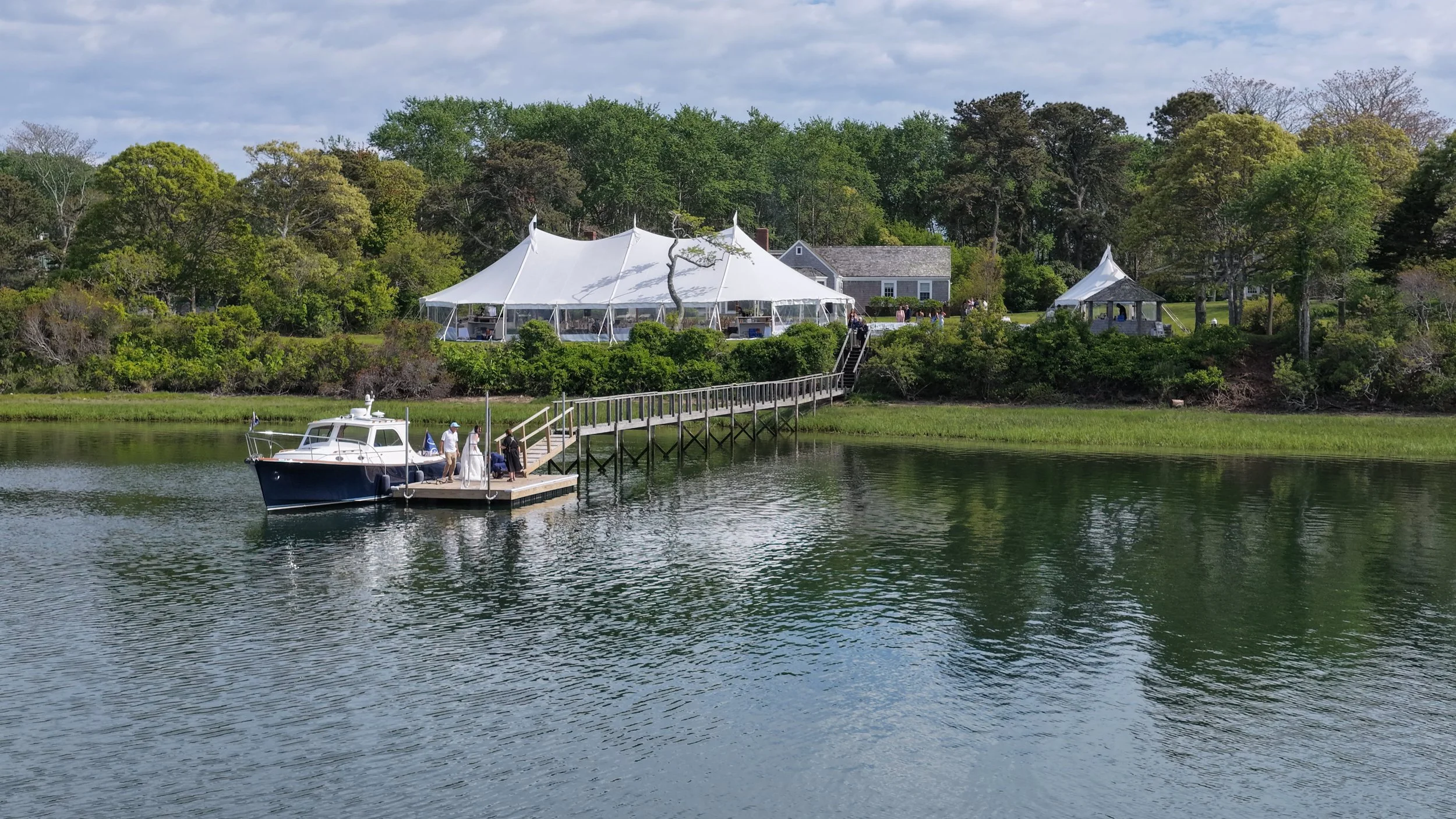 A waterfront wedding scene showing a boat docked at a pier, a bride and groom in wedding attire, and a large white tent set up on a lush green island with trees and a small building in the background.