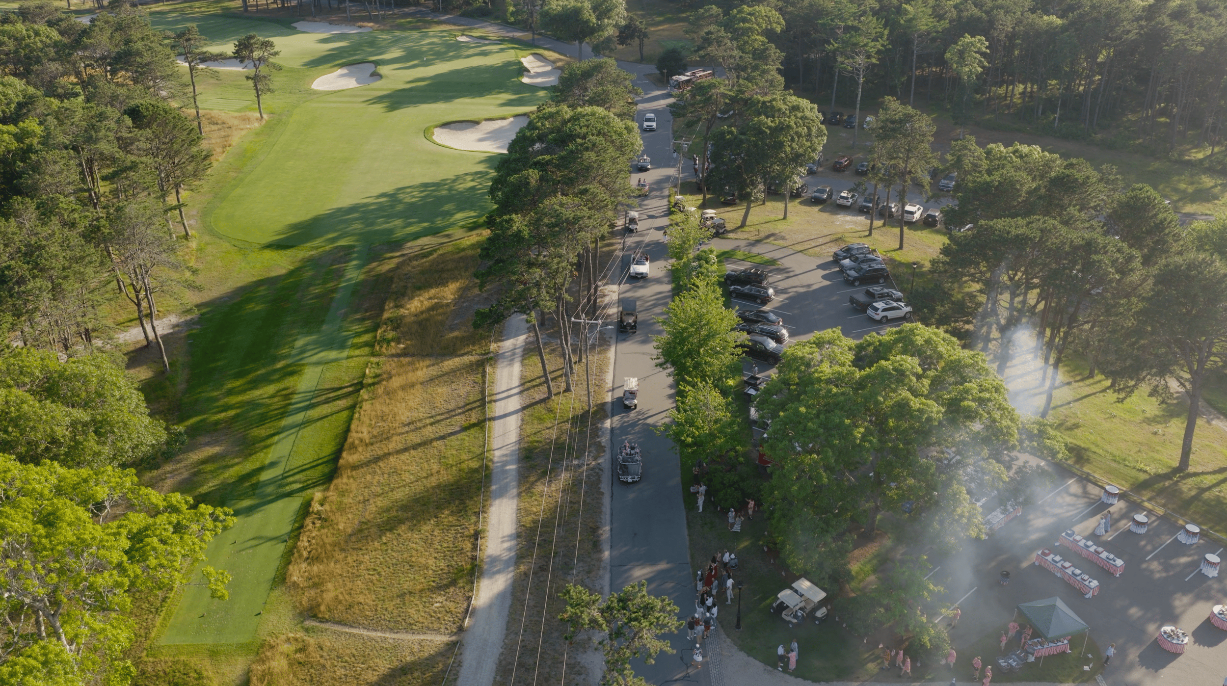 An aerial view of a golf course with green fairways, sand traps, surrounded by trees, and a parking lot with parked cars and some people.