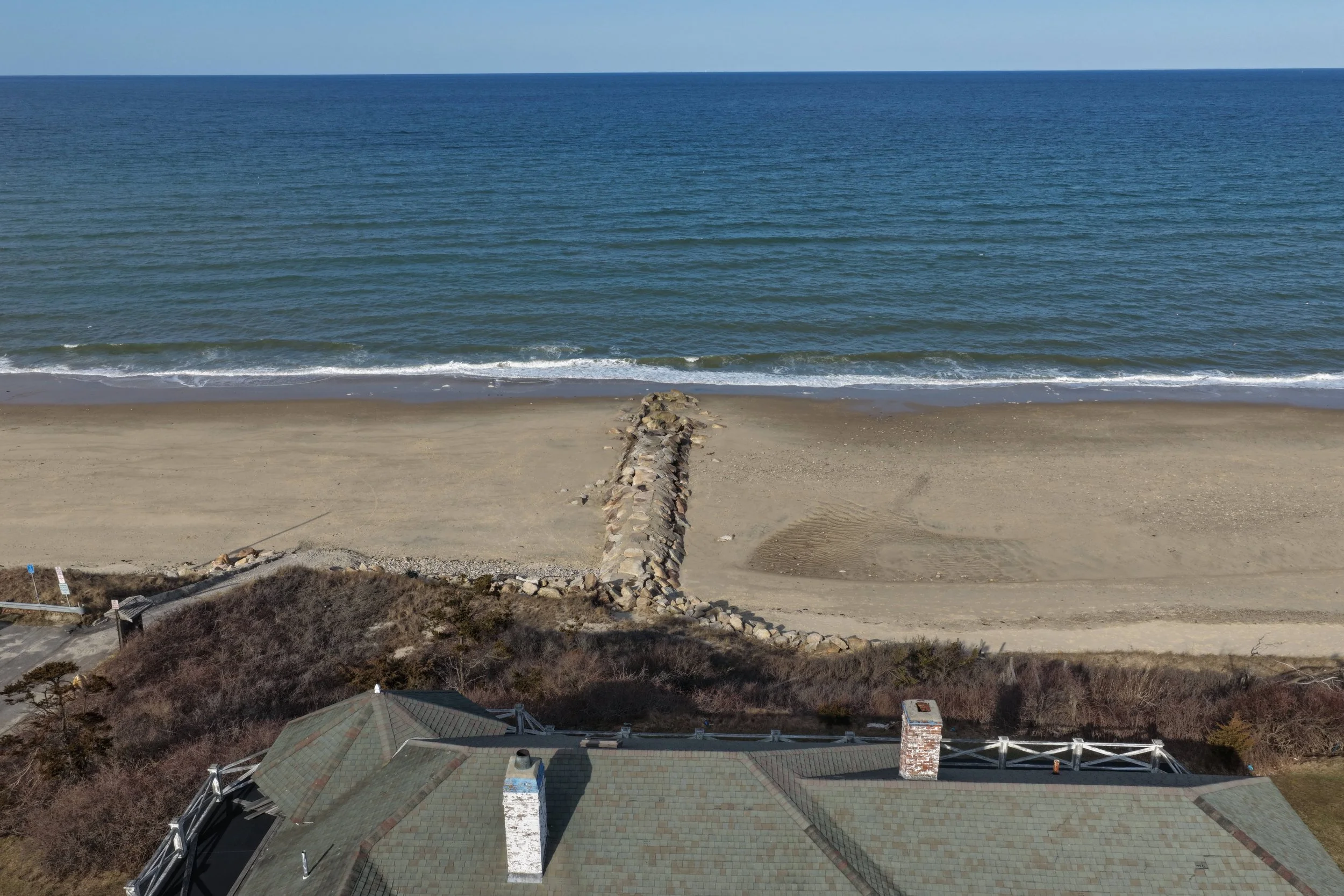 Aerial view of a beach with a stone jetty extending into the ocean, sandy shore, and a house with a green roof in the foreground.