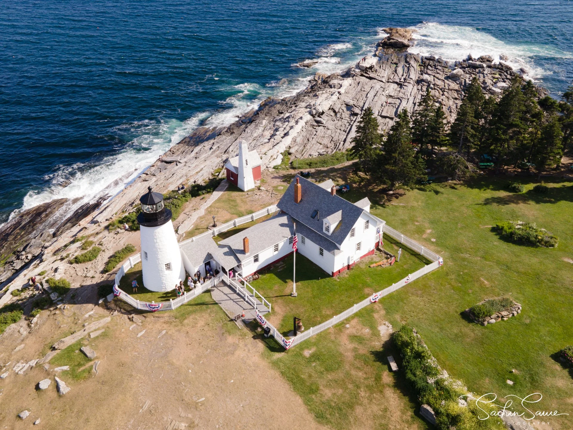 Aerial view of a white lighthouse, a keeper's house, and additional small buildings on a rocky coastline with trees, grass, and ocean waves.