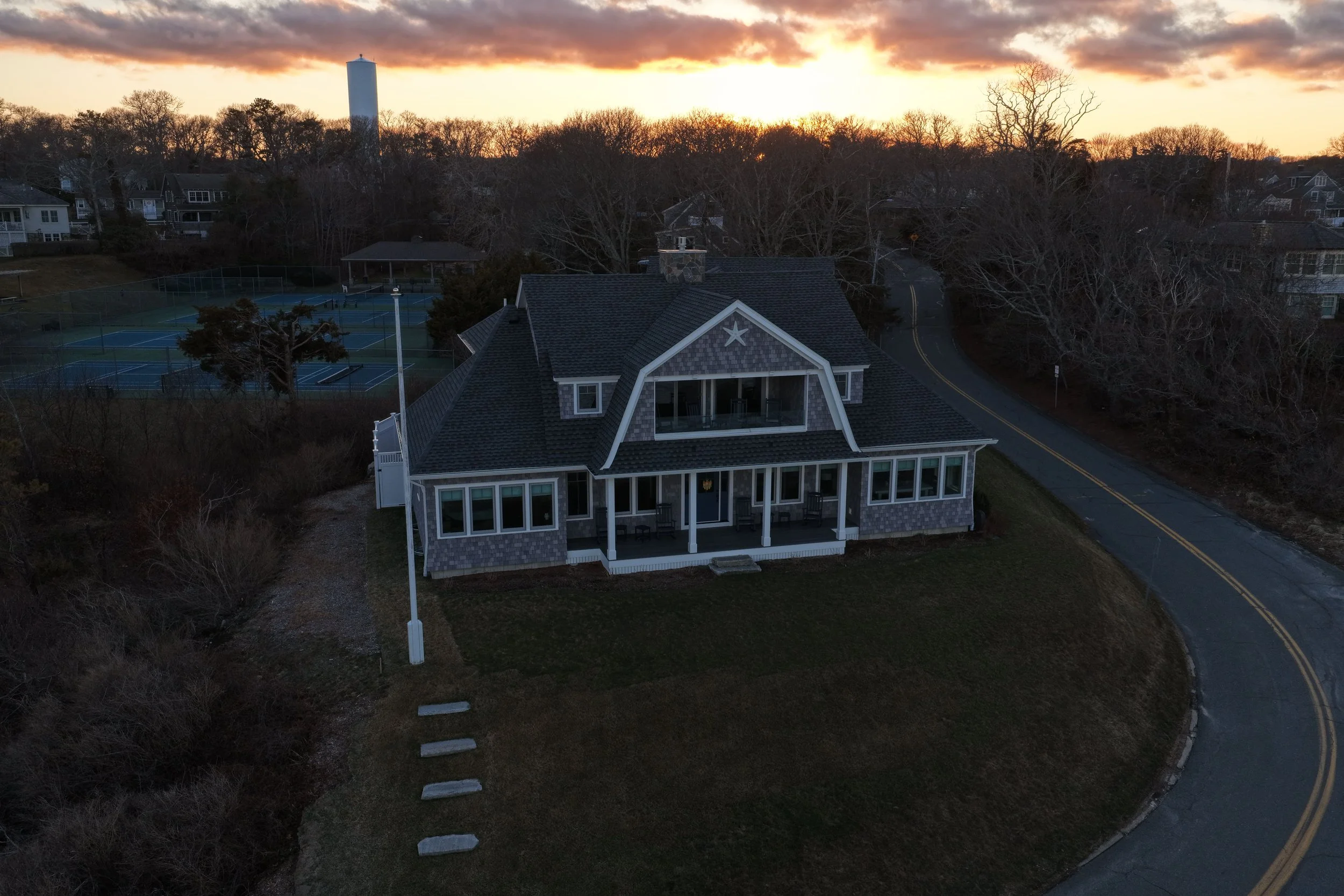 A large two-story house with a wraparound porch, multiple windows, and a steep gabled roof, situated on a curved road with leafless trees around it. Tennis courts are visible behind the house, and the sky shows a sunset with orange and purple hues.