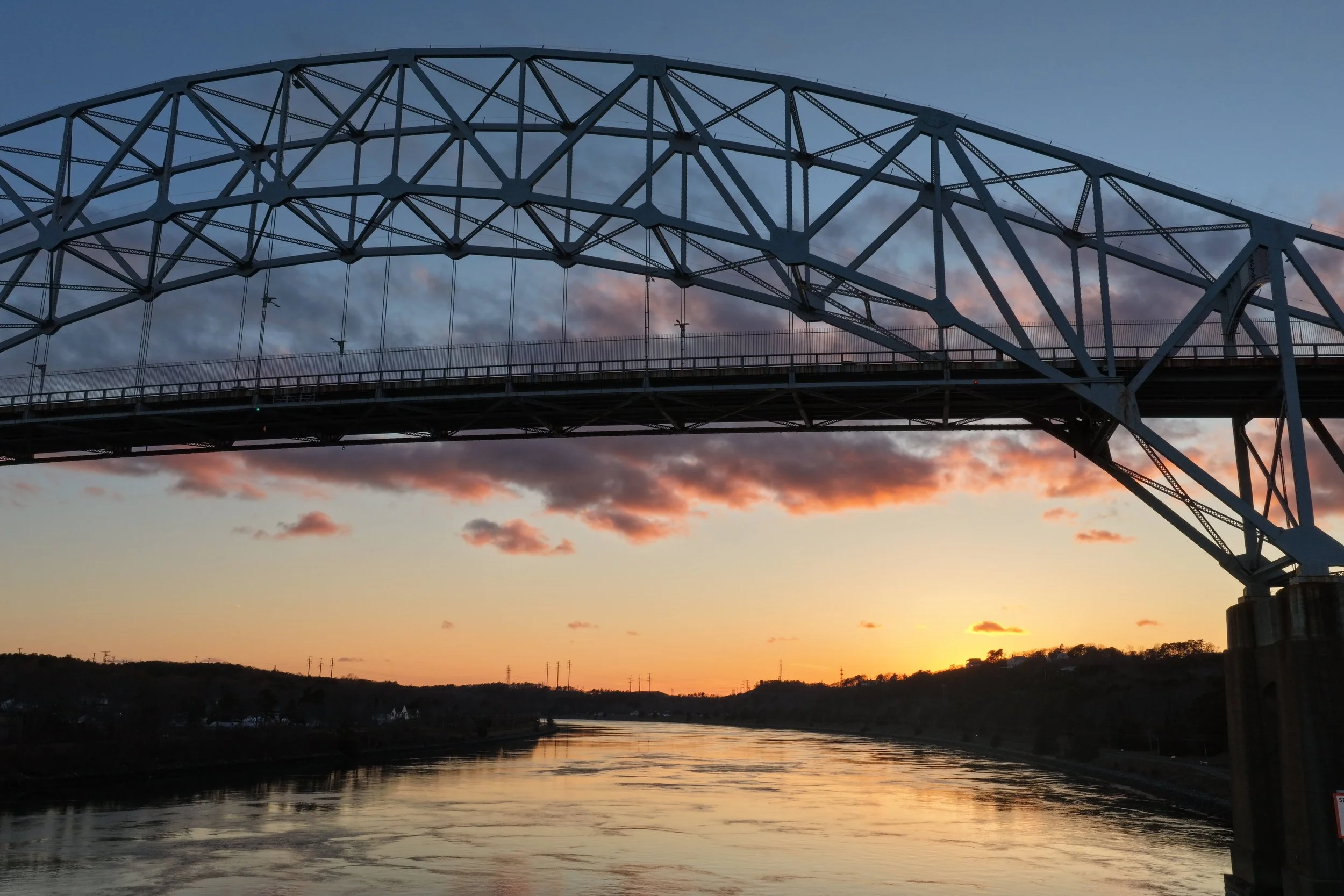 Photo of a large steel arch bridge over a river during sunset, with pink and orange clouds in the sky and water reflecting the colors.