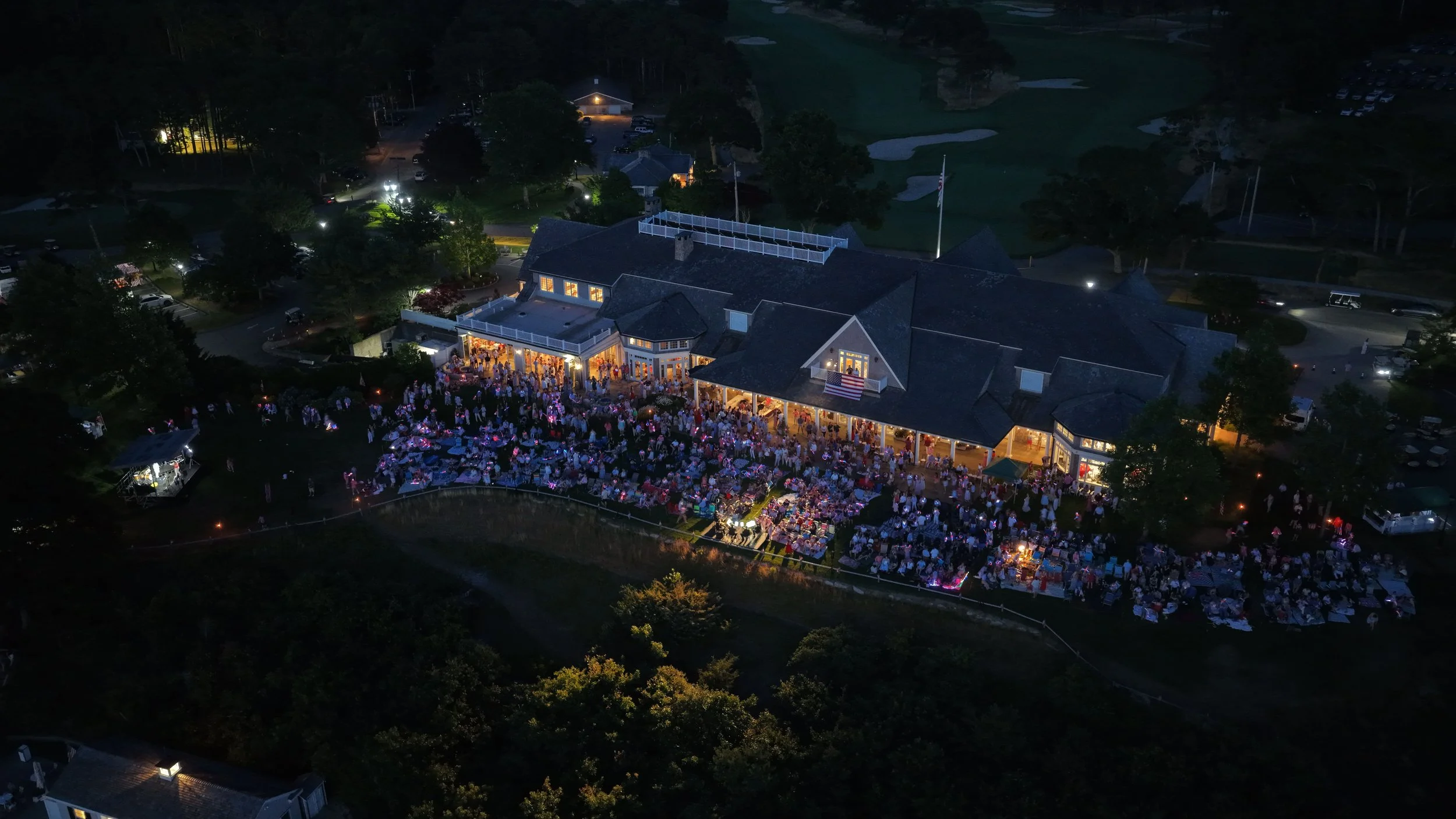 An aerial night view of a large outdoor party with many people gathered around tables outside a building with lights, set in a green landscape with trees, parking lots, and a golf course in the background.