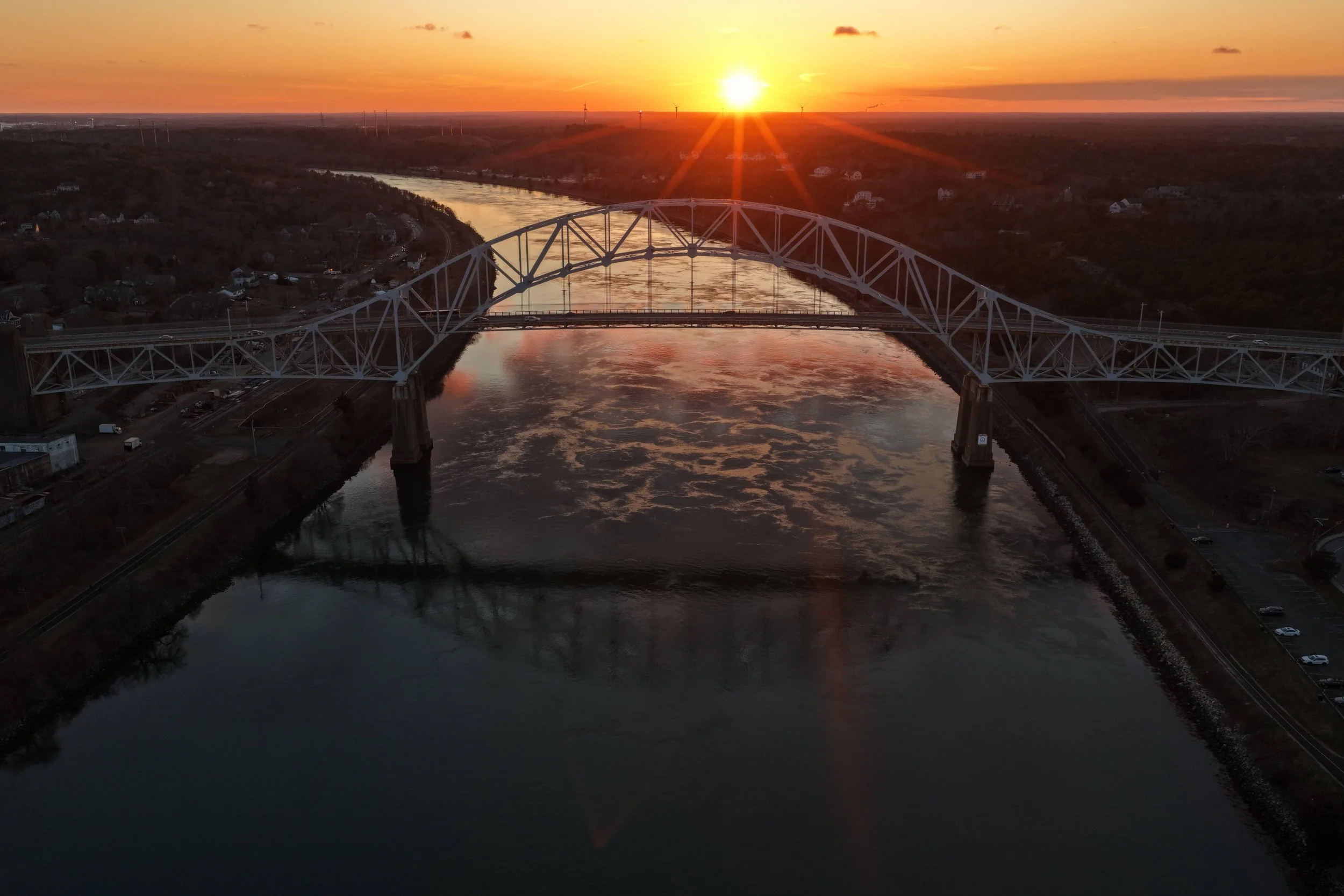 A bridge over a river at sunset with a clear sky and the sun just above the horizon, reflecting on the water.