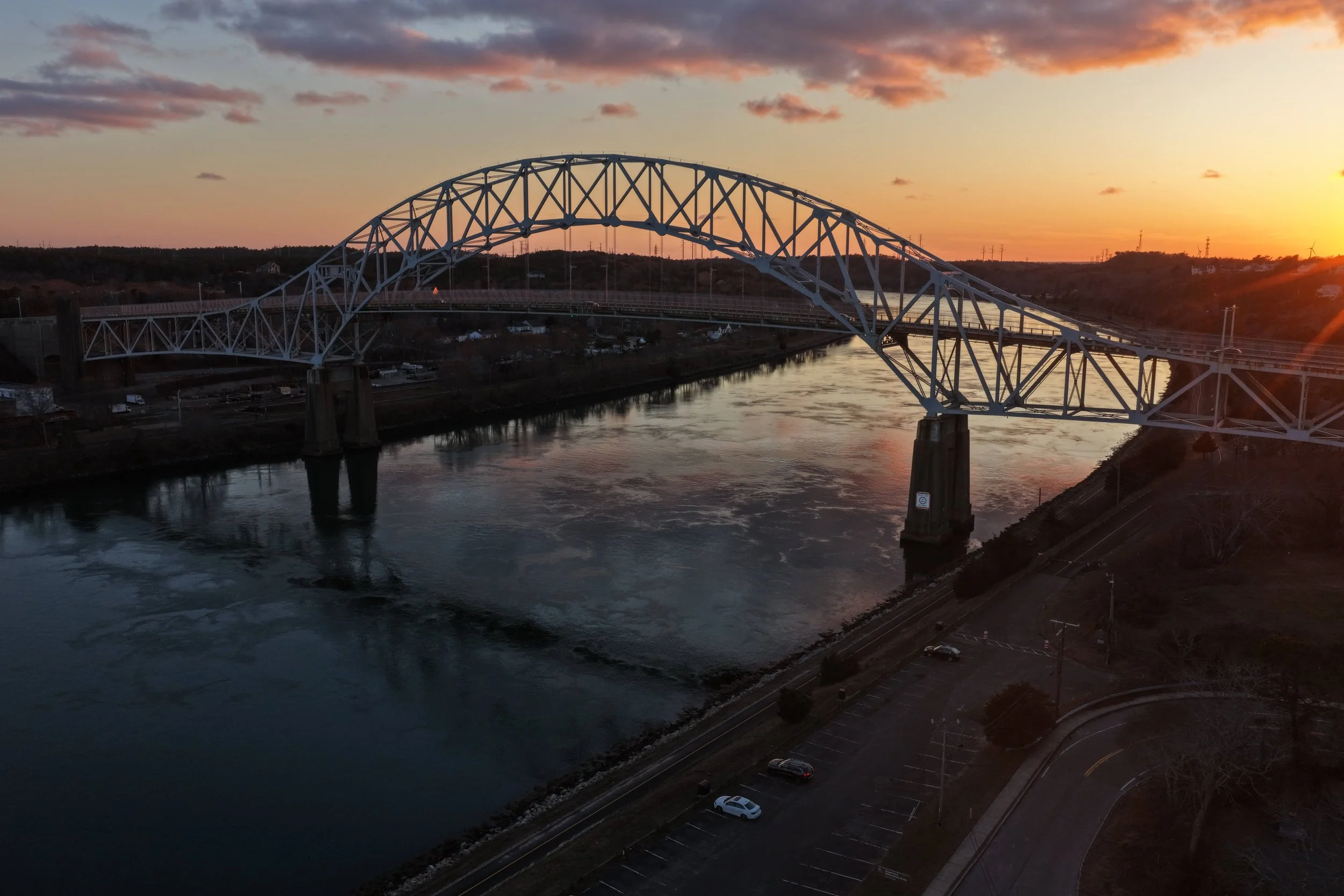 A bridge spanning a river at sunset with a partly cloudy sky and parked cars below.