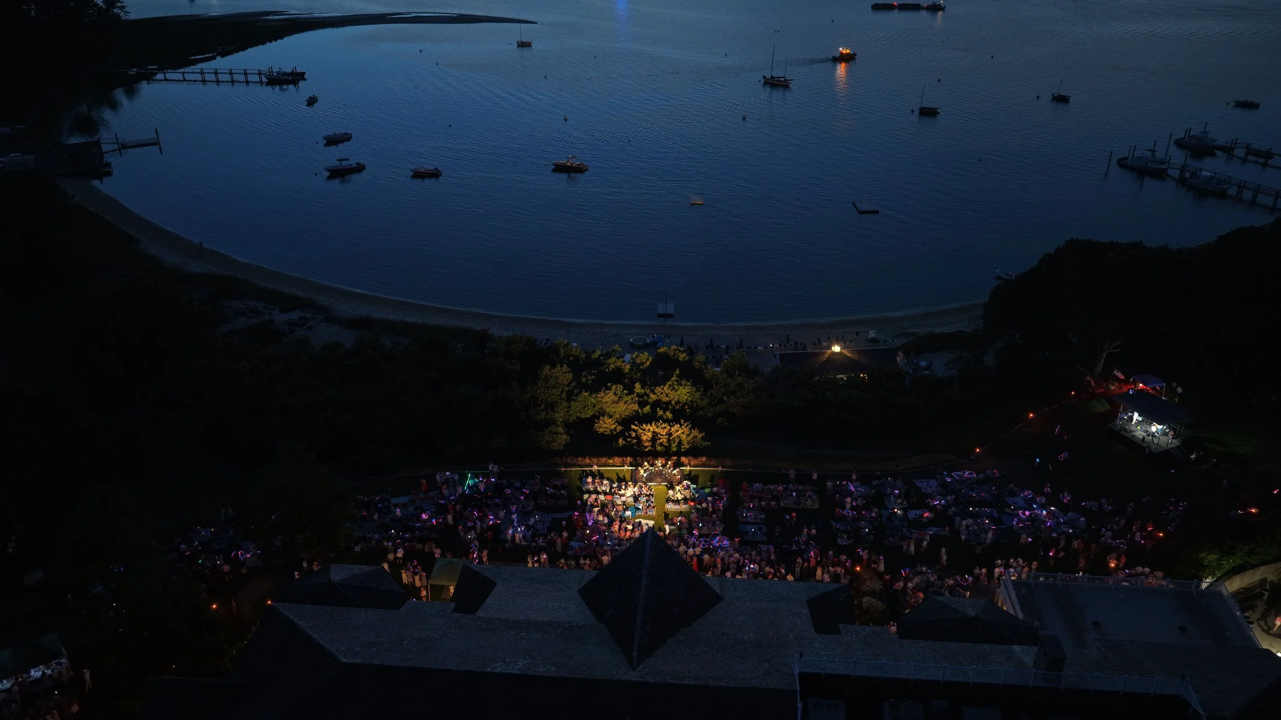 Aerial night view of a marina with boats docked in the water, a beach area with tents and people, and a waterfront scene illuminated by colorful lights.