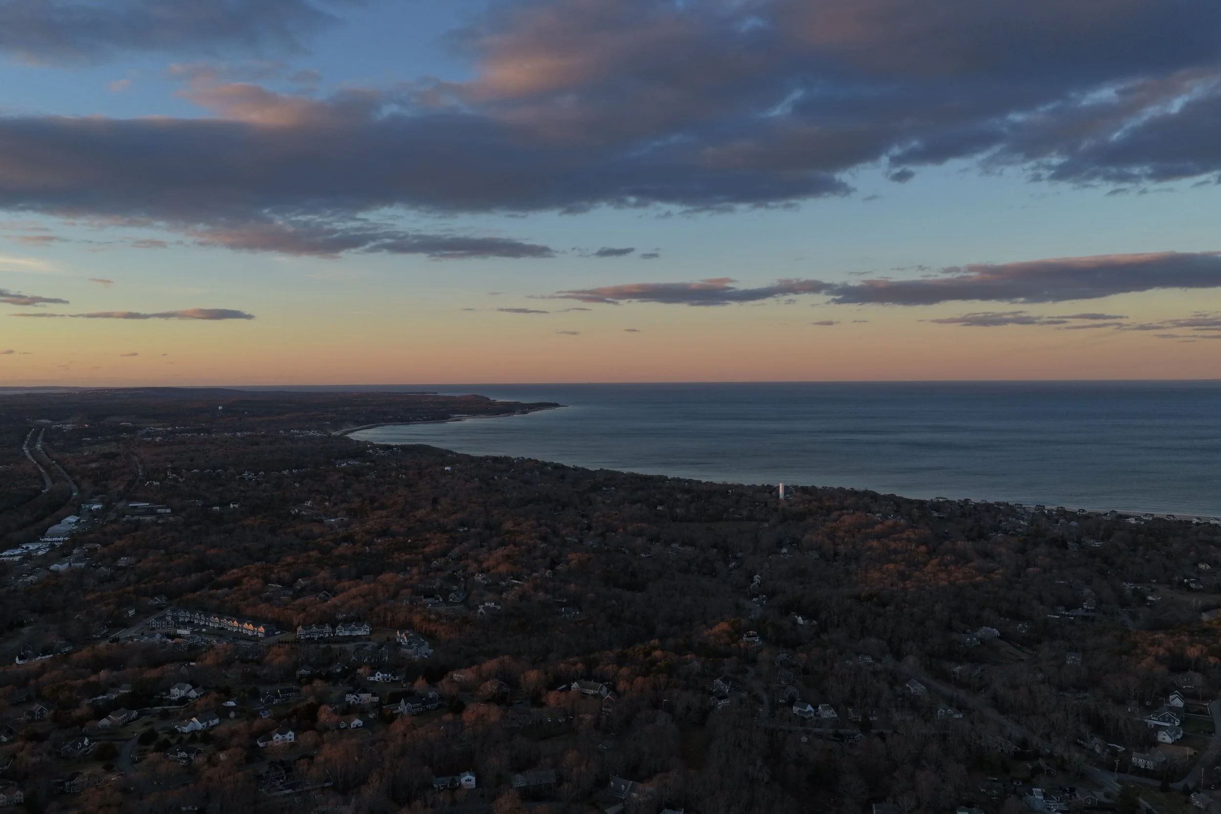 Aerial view of a coastal landscape during sunset, showing the ocean, shoreline, and residential area with houses and roads surrounded by trees.