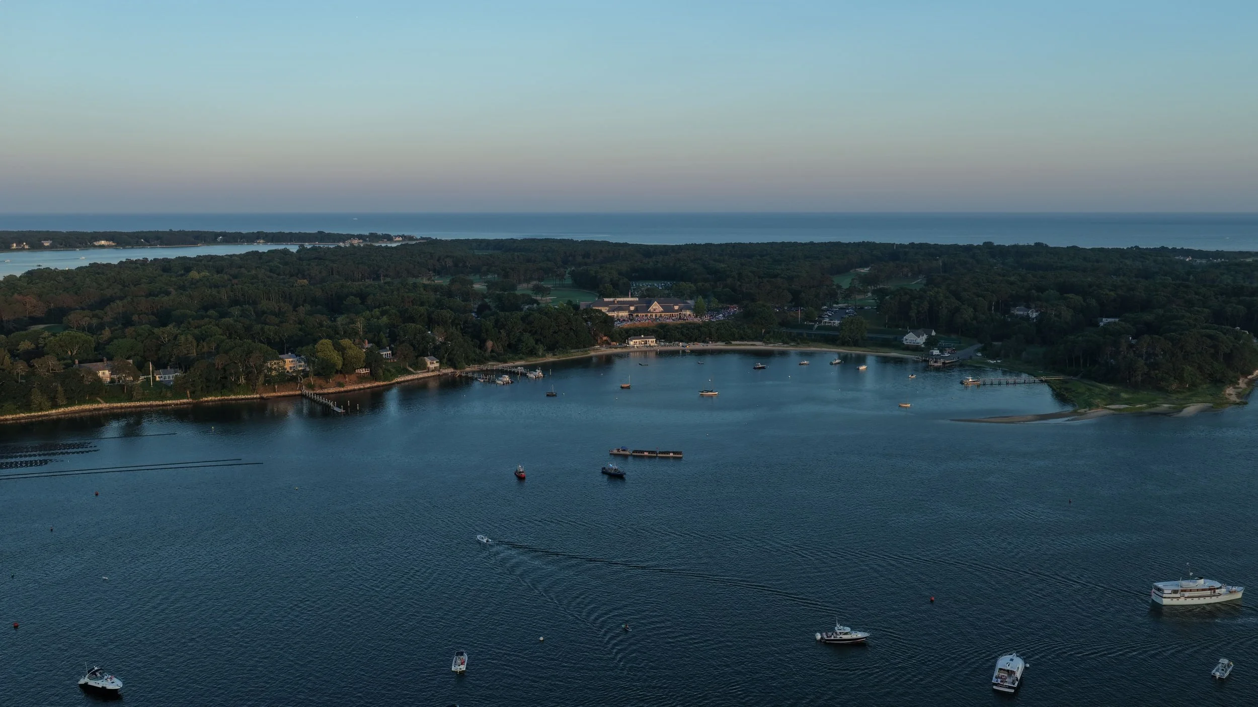 An aerial view of a coastal area with a bay filled with boats, a small harbor, green trees, and houses along the shoreline. The background shows a distant coastline and open ocean under a clear sky.