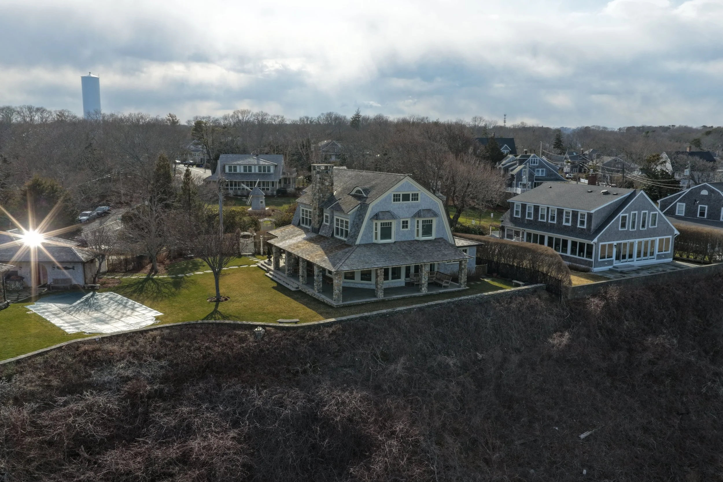 A large two-story house with a wraparound porch in a residential neighborhood, surrounded by trees and other houses, with a hillside in the foreground and a cloudy sky above.