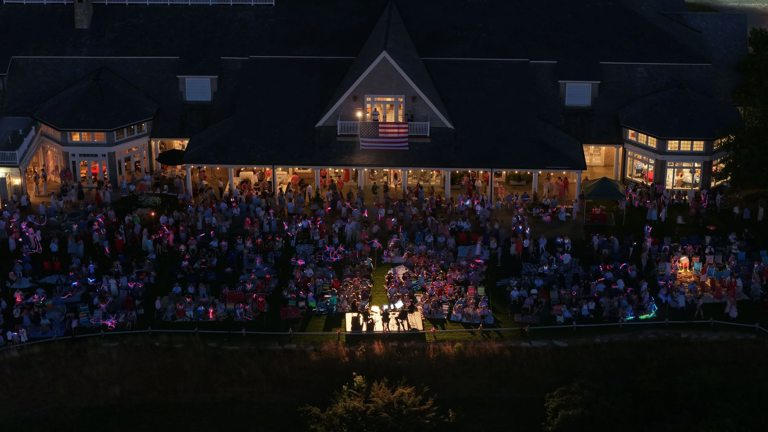 Nighttime outdoor gathering at a large house with illuminated windows, a central balcony with an American flag, and a crowd of people with glowing accessories.