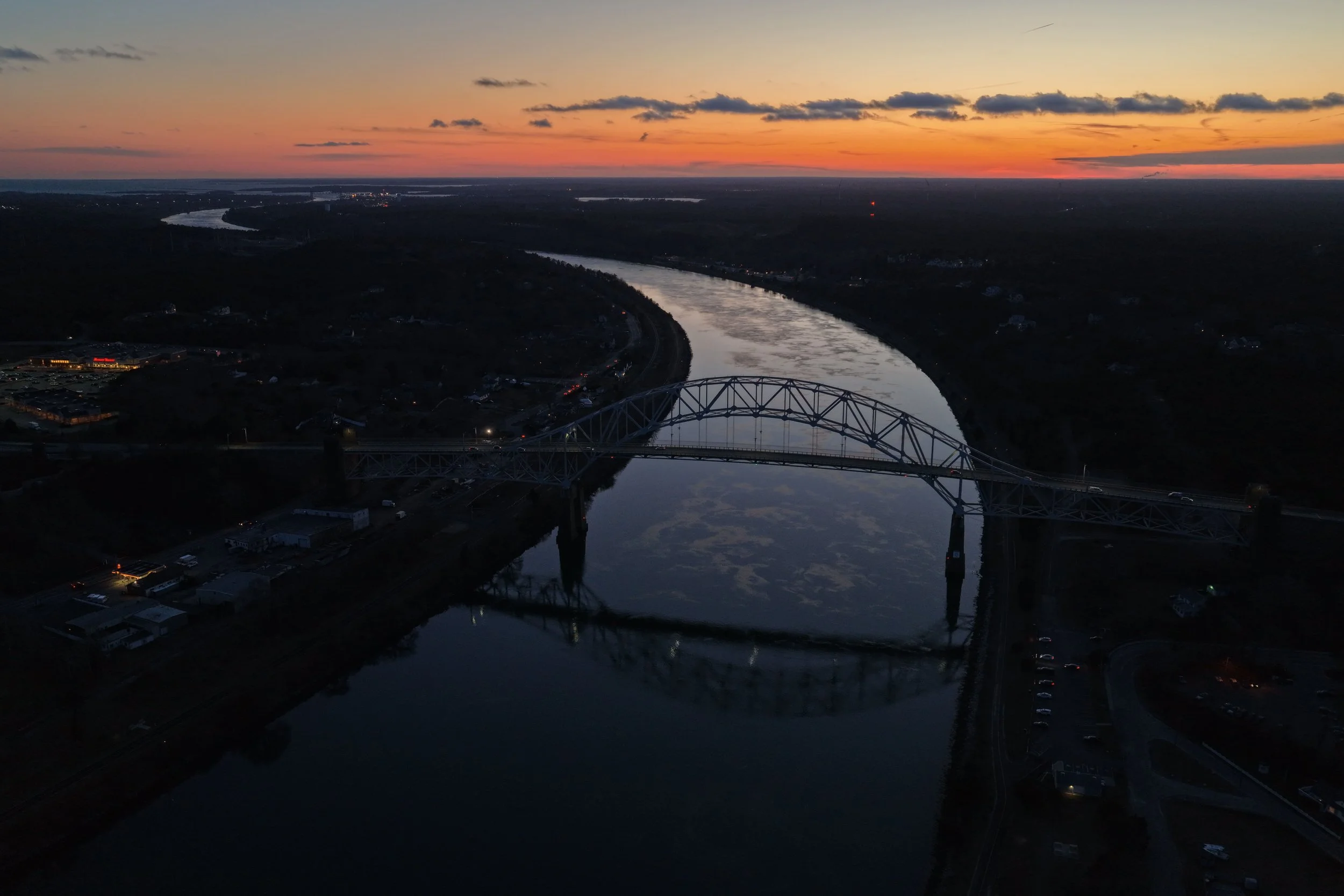 Aerial view of a river at sunset with a bridge crossing over it, with the sky displaying orange, pink, and purple hues and a reflection of the sky on the water.