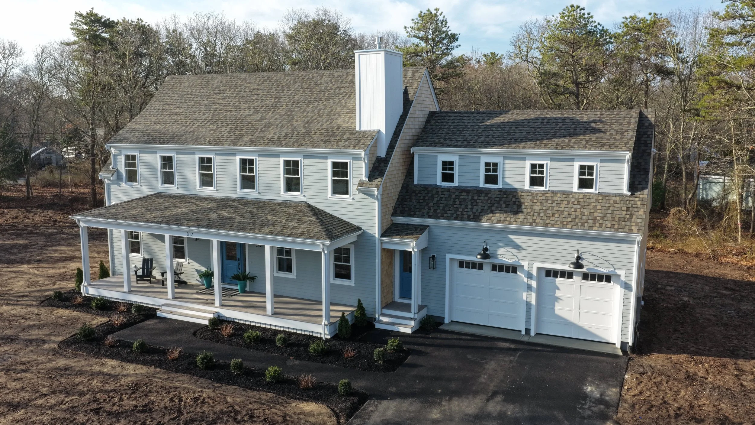 A modern two-story house with a porch, multiple windows, a garage, and a pitched roof, surrounded by a bare yard and trees in the background.