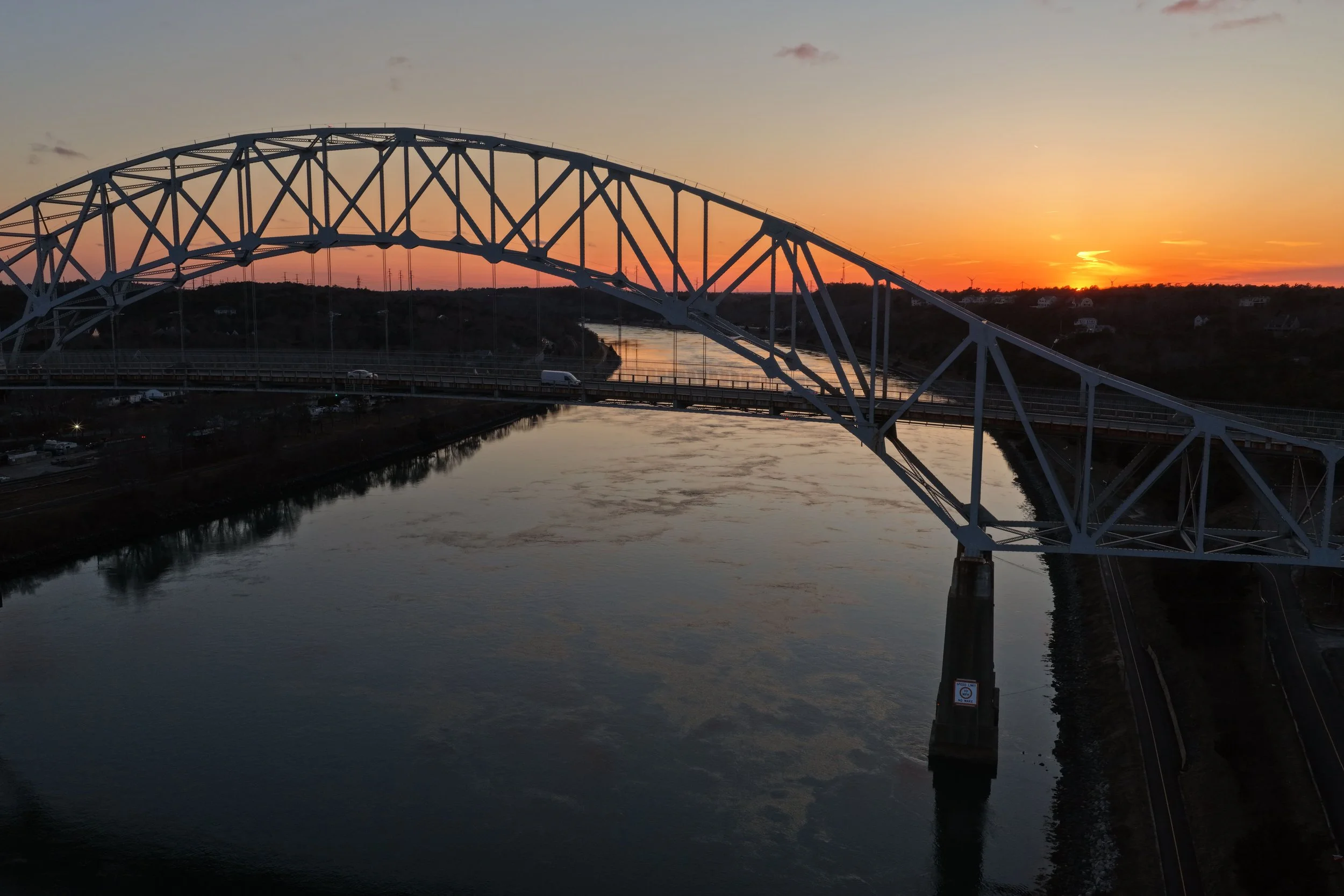 A bridge over a river during sunset with an orange sky and a few clouds.