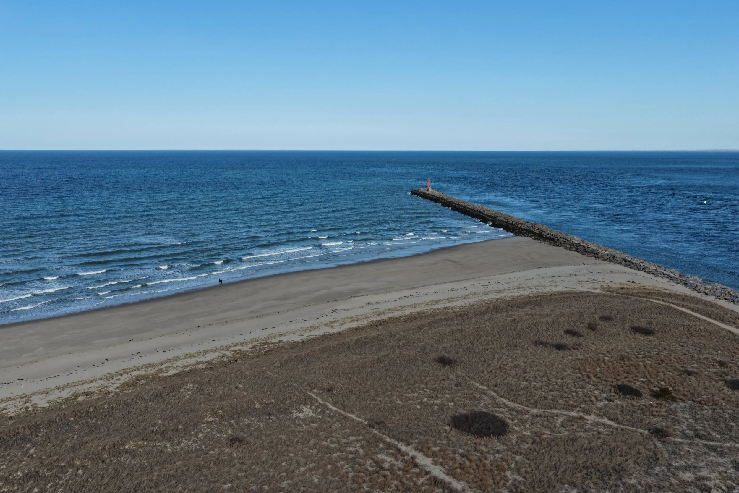A beach with light sand and sparse vegetation, a long rocky pier extending into the ocean, with a lighthouse at the end. The water is calm with small waves, and the sky is clear and blue.