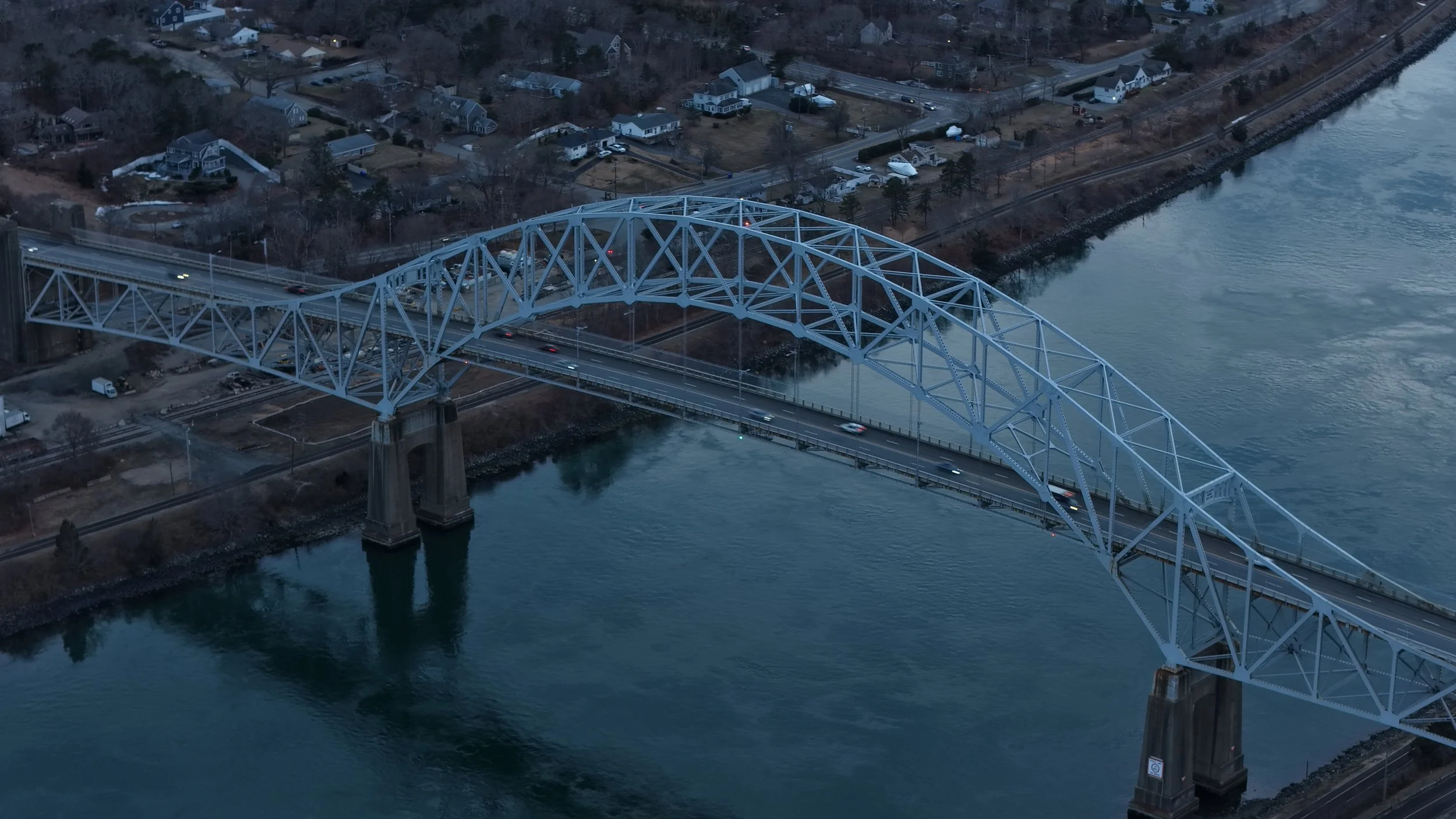 An aerial view of a large blue steel arch bridge crossing a body of water, with a residential area in the background.