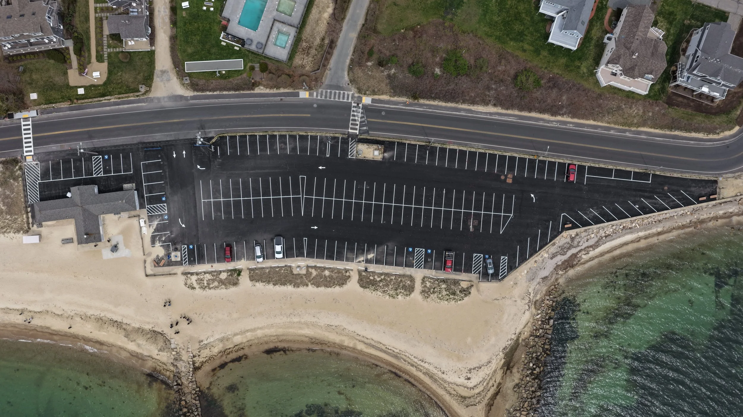 An aerial view shows a beachfront parking lot with multiple empty parking spaces, a small row of cars parked, and a road leading to houses with swimming pools and apartments. The image also captures a sandy beach and a section of ocean with rocks and green water.