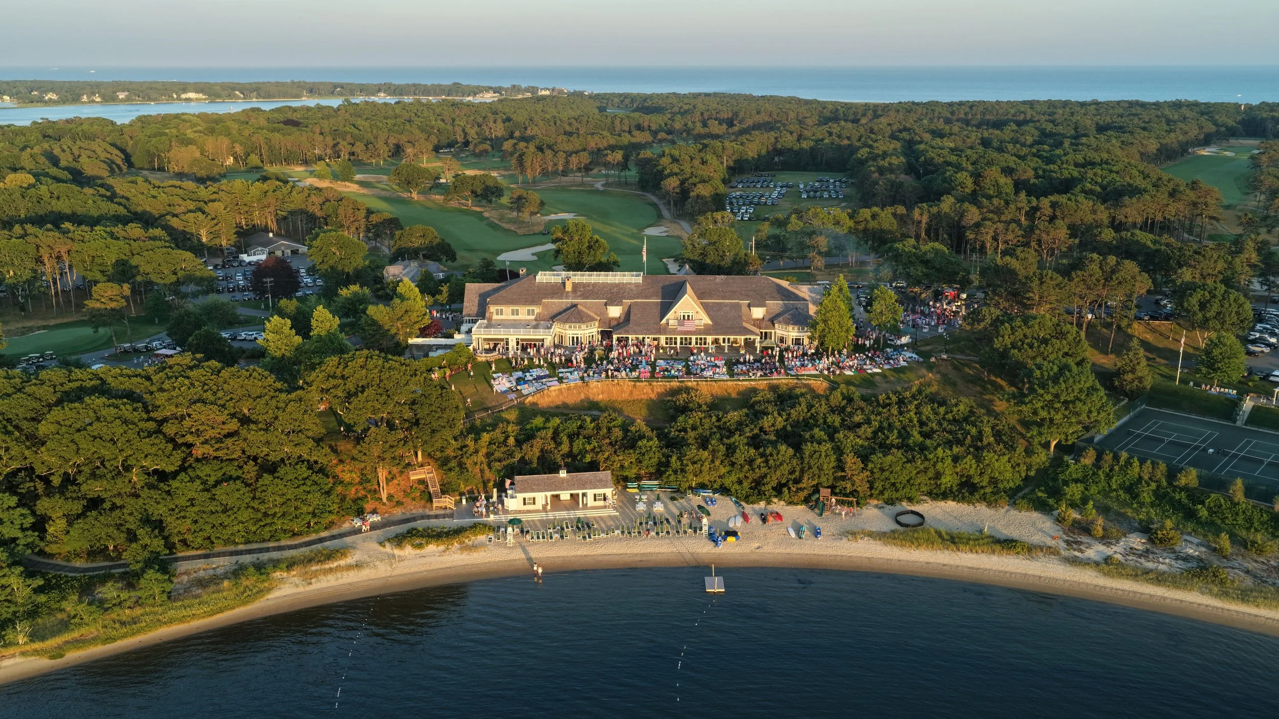 Aerial view of a waterfront restaurant with an outdoor seating area, surrounded by a forested golf course and parking lot, near a beach and ocean in the background.