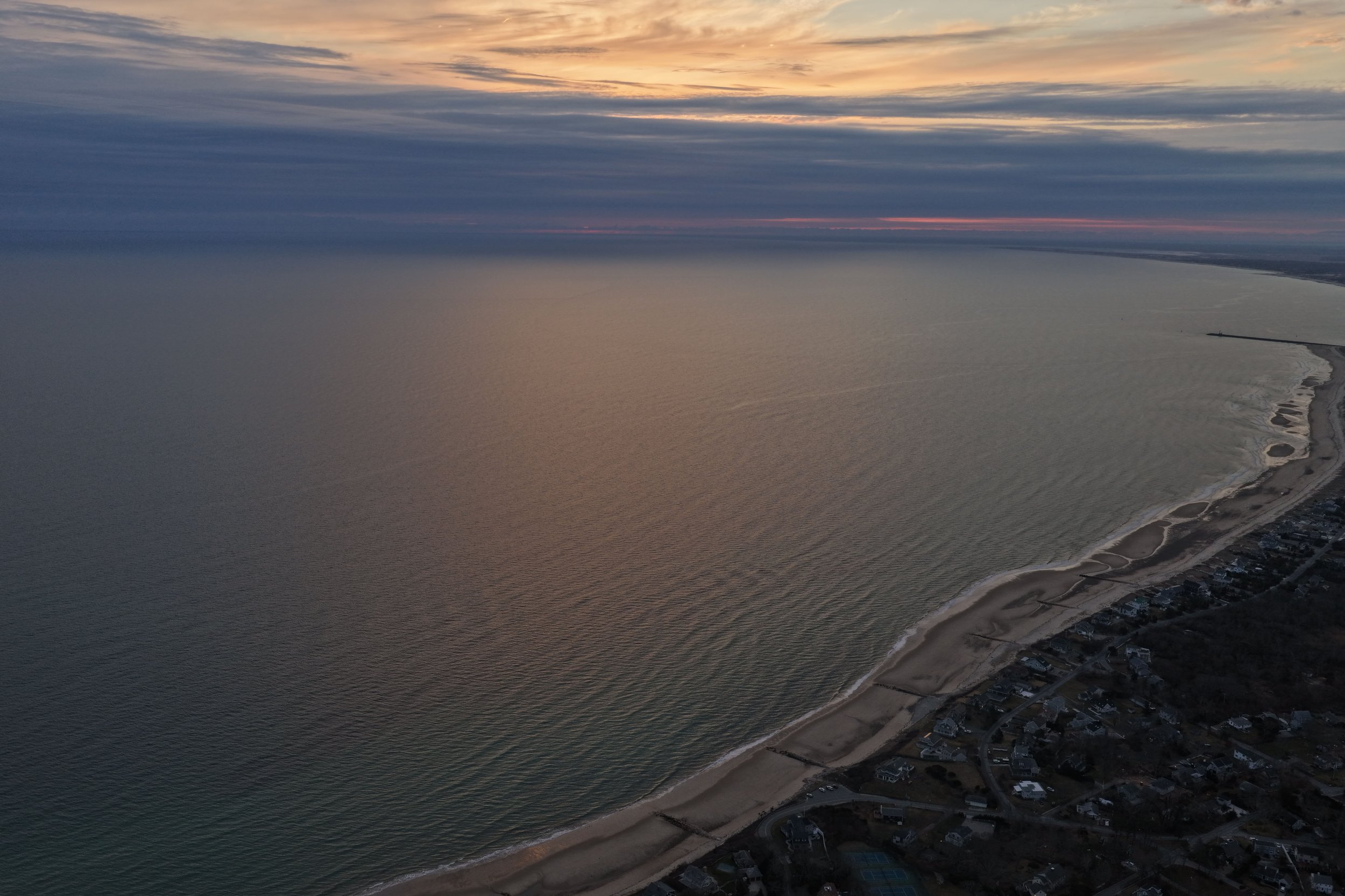 Aerial view of a beach at sunset with calm water, sandy shoreline, and houses along the coast.