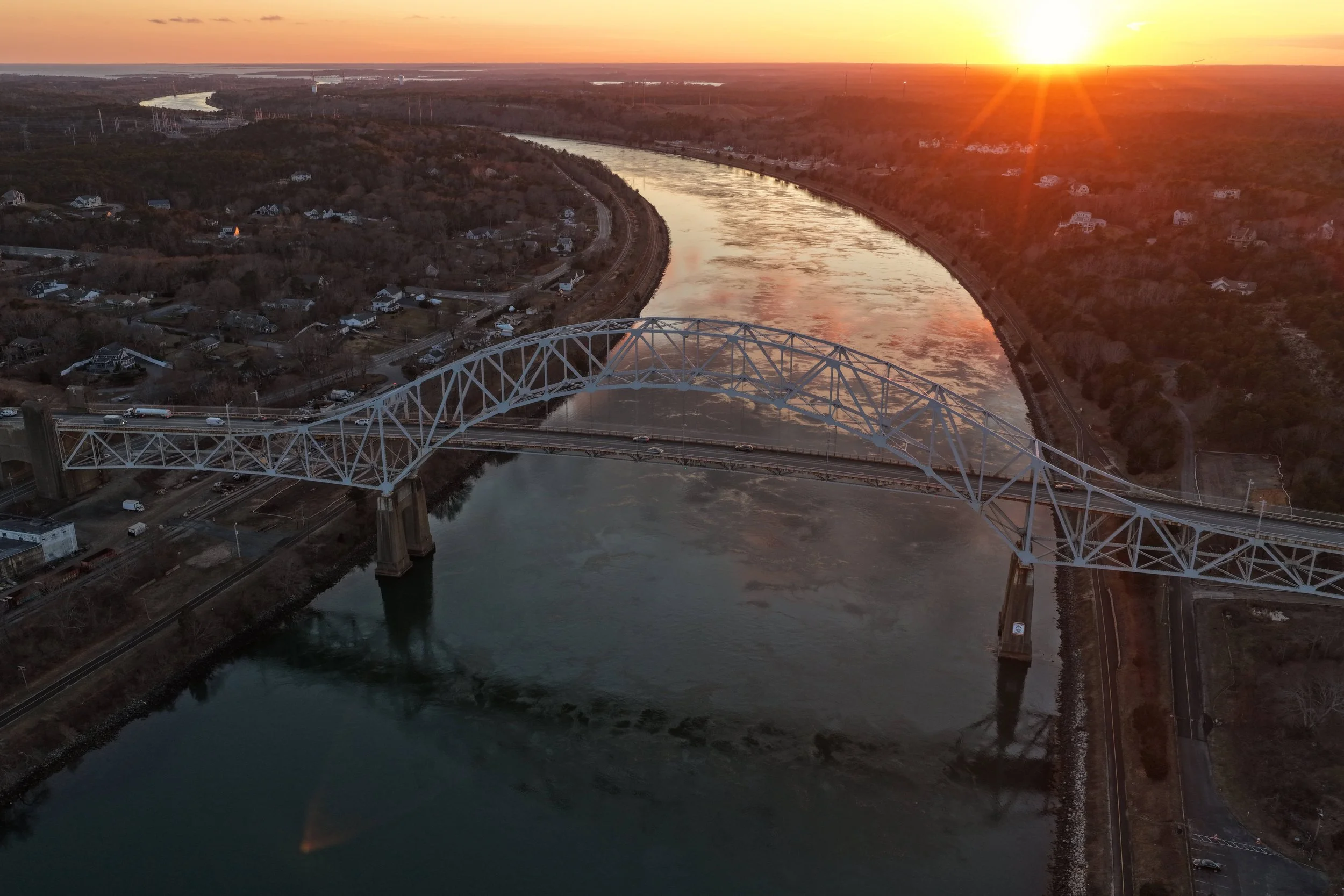 An aerial view of a large steel arch bridge spanning a wide river during sunset, with houses and trees on both sides of the riverbank.
