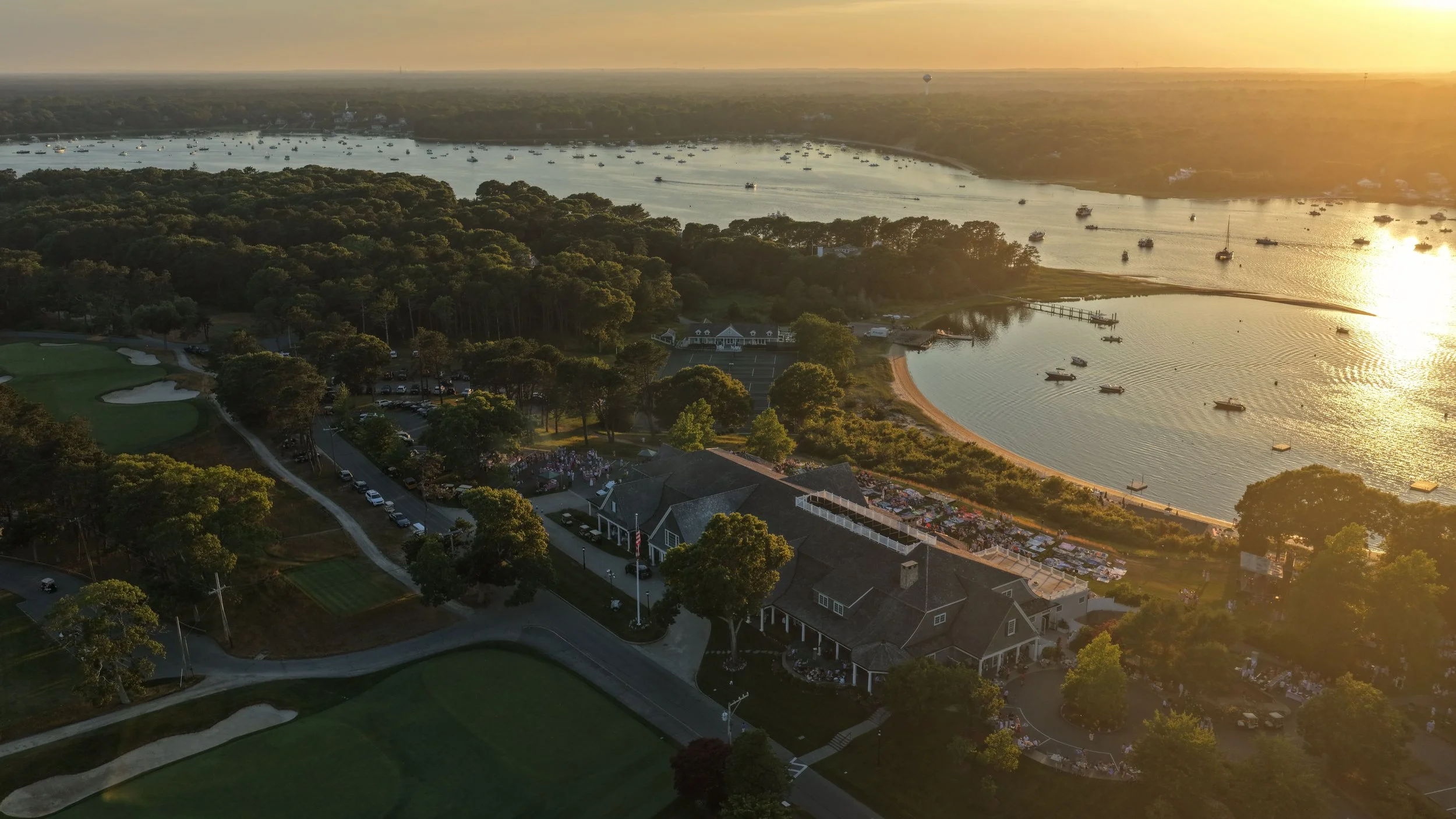 Aerial view of a coastal area at sunset showing a harbor with boats, a golf course, and a building with outdoor seating. The sun reflects on the water, and the scene includes lush green trees and residential houses.