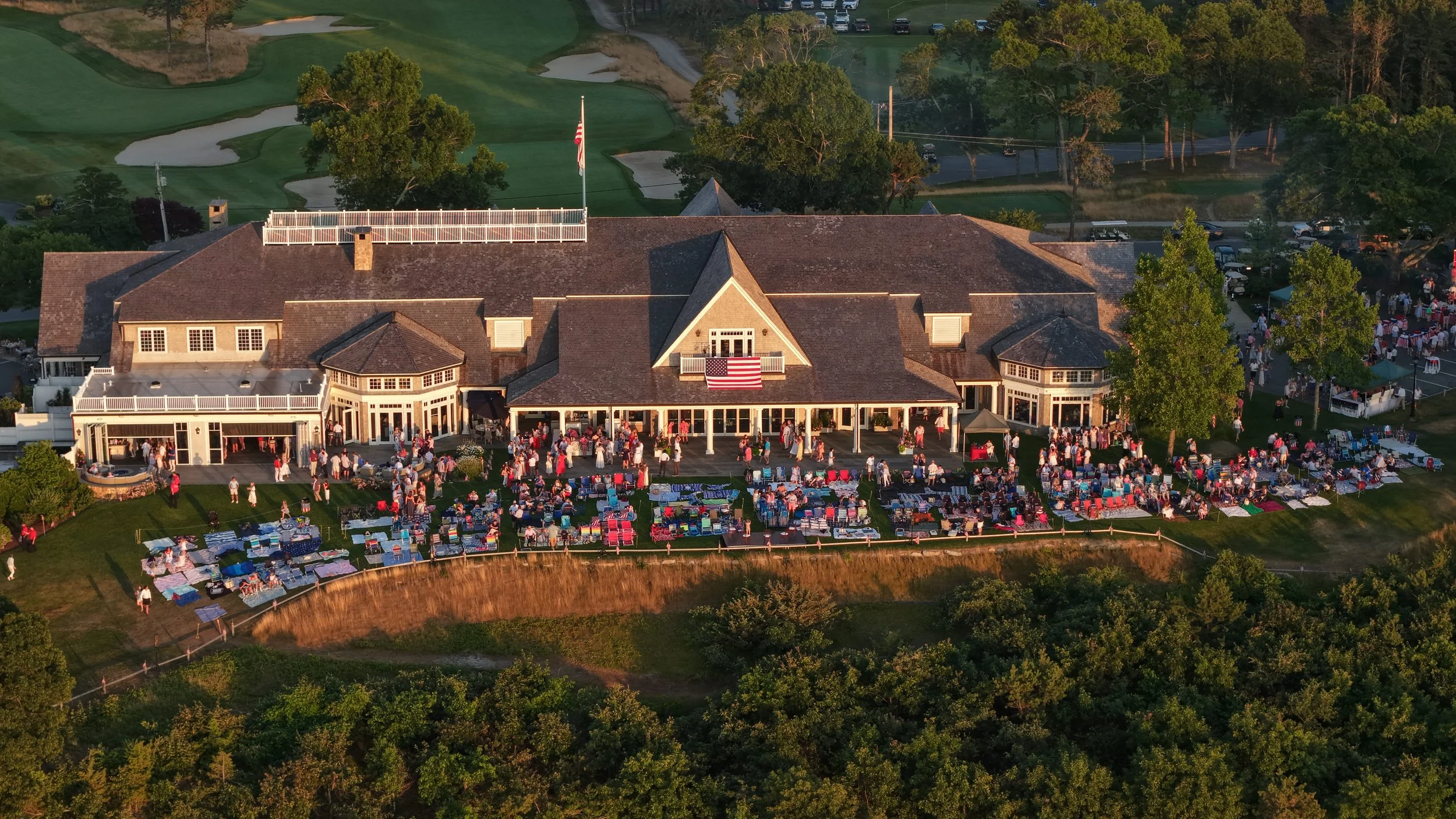 An aerial view of a building with many guests gathered outside for an event, decorated with American flags, with a golf course in the background.