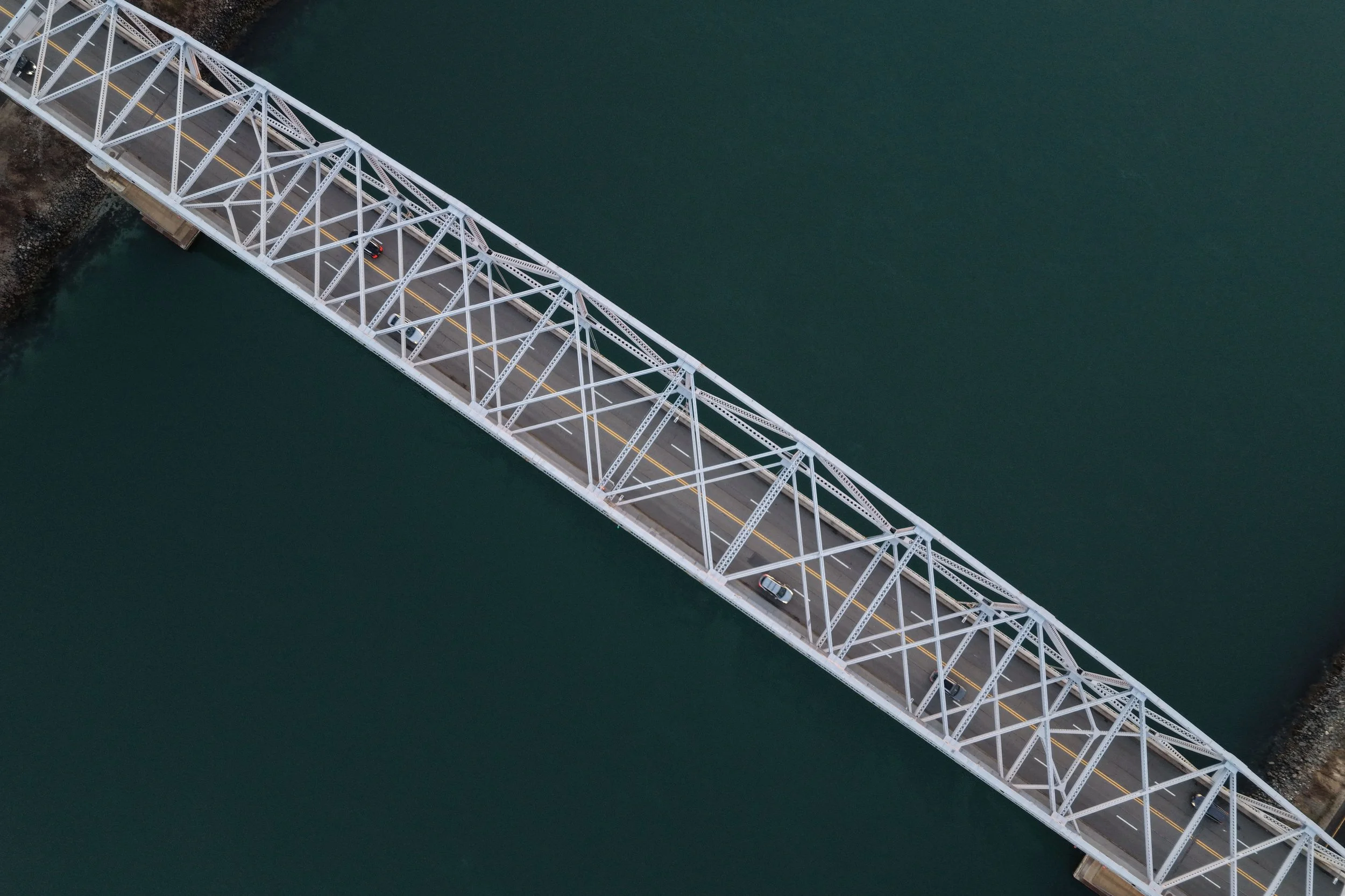 Aerial view of a white metal truss bridge crossing over a body of water with a few cars on it.