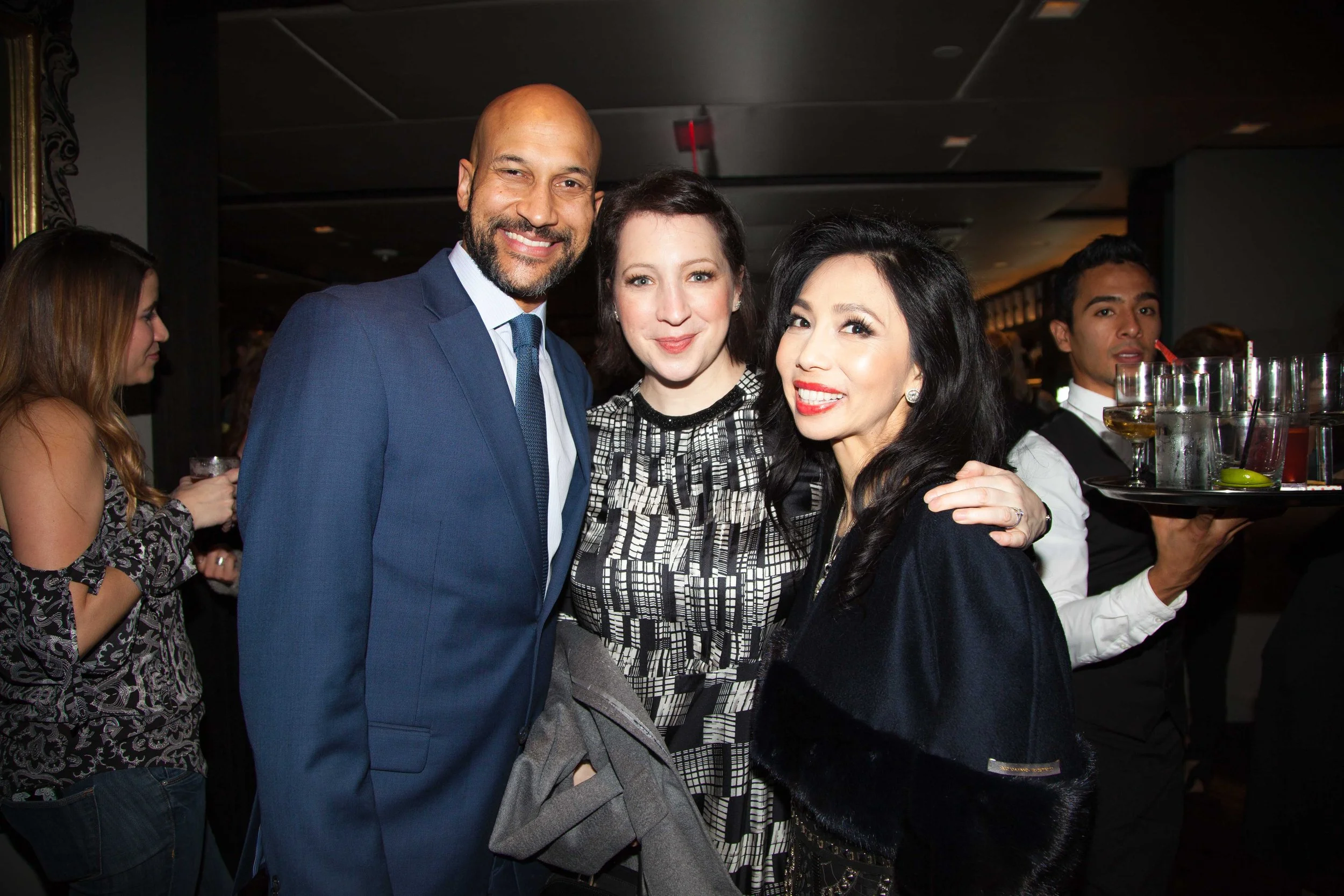 Three people smiling and posing for a photo at a social event, with a woman holding a tray of drinks in the background.