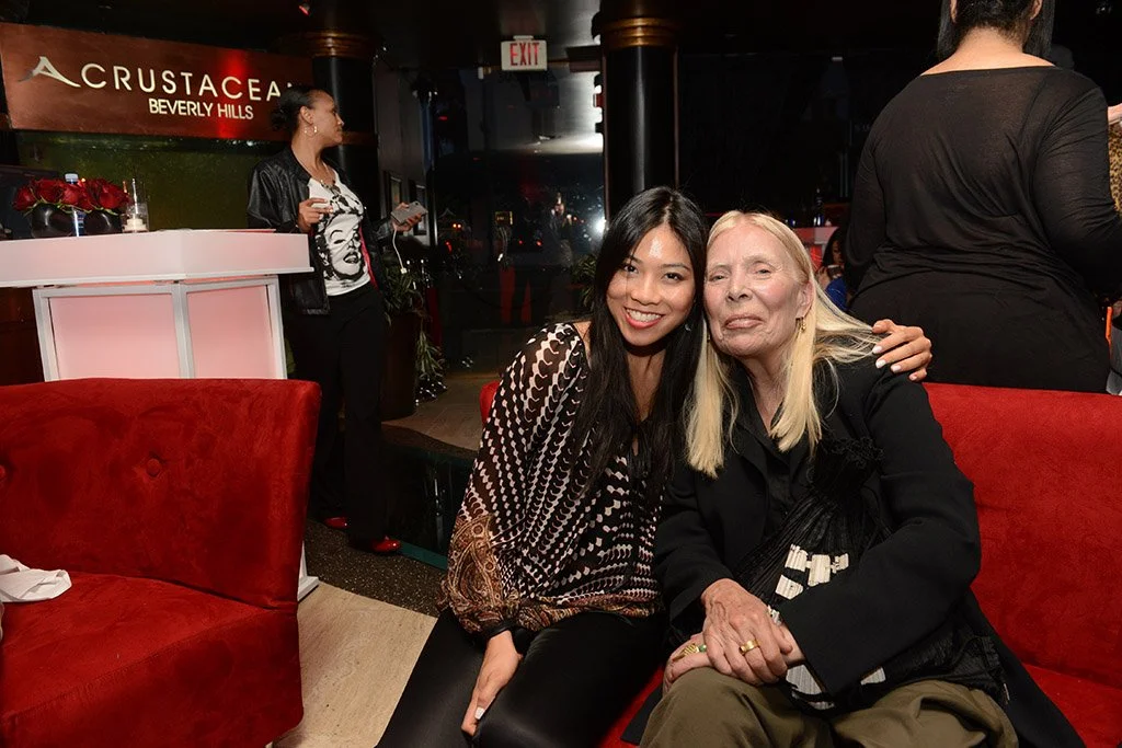 Two women sitting on a red sofa in a restaurant, smiling and posing for the photo. There is a woman in the background near a counter, holding a notebook. The restaurant has a sign that reads "CRUSTACEAN BEVERLY HILLS."