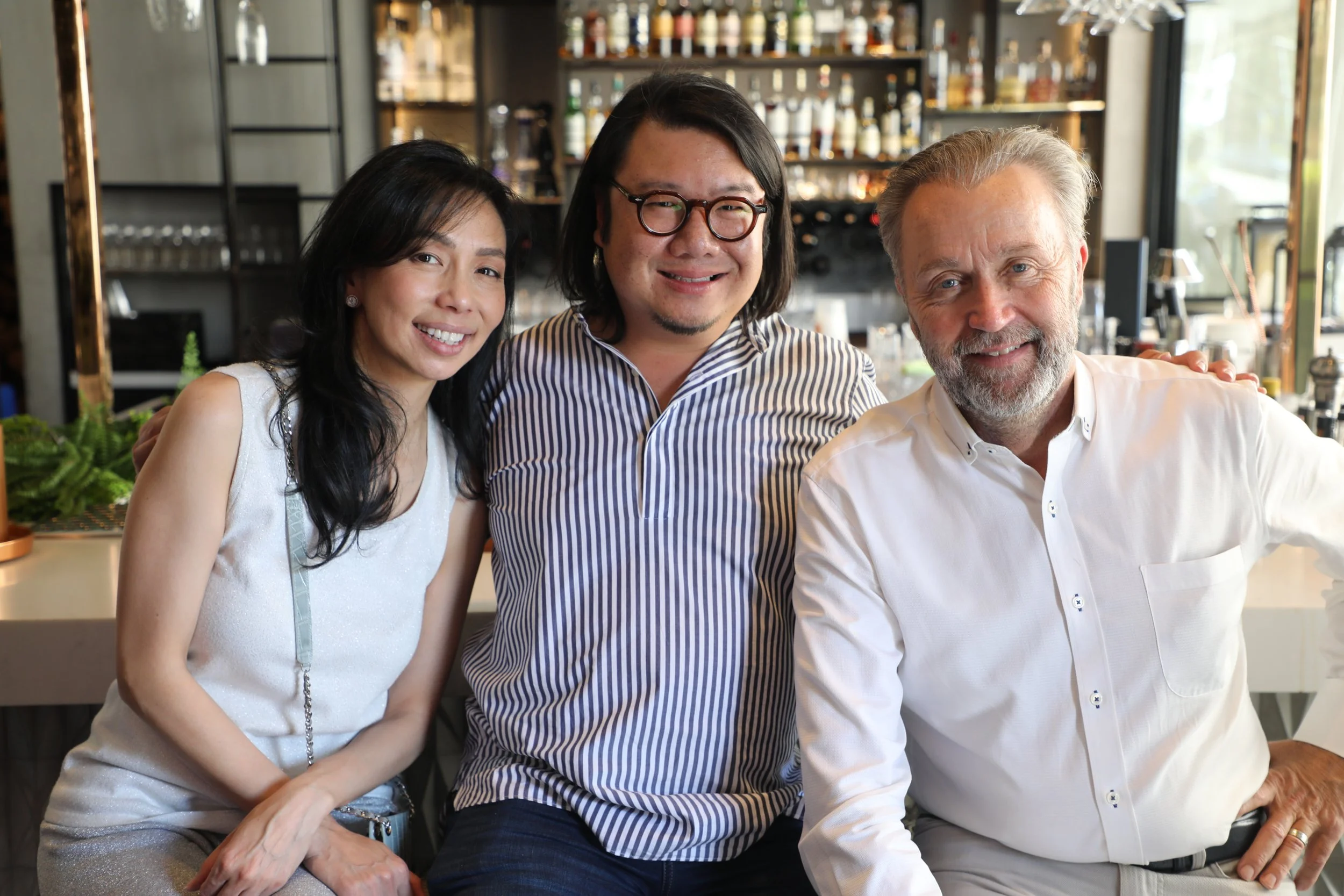 Three friends smiling at a bar or restaurant, sitting close together.