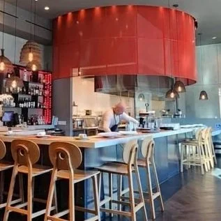 A cozy indoor dining area with a small round table, two wooden chairs, a decorative bonsai-like plant in a pot, a lit fireplace with a modern rectangular opening, and a stand holding wine bottles and glasses, all against a textured brown wall.